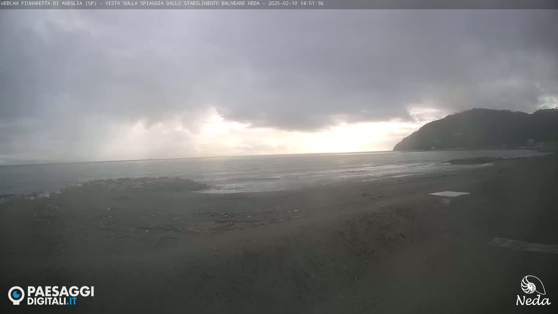 A dark, overcast day shows a beach with a dark-grey sandy shore, small waves lapping at the coast, and a hill in the background.