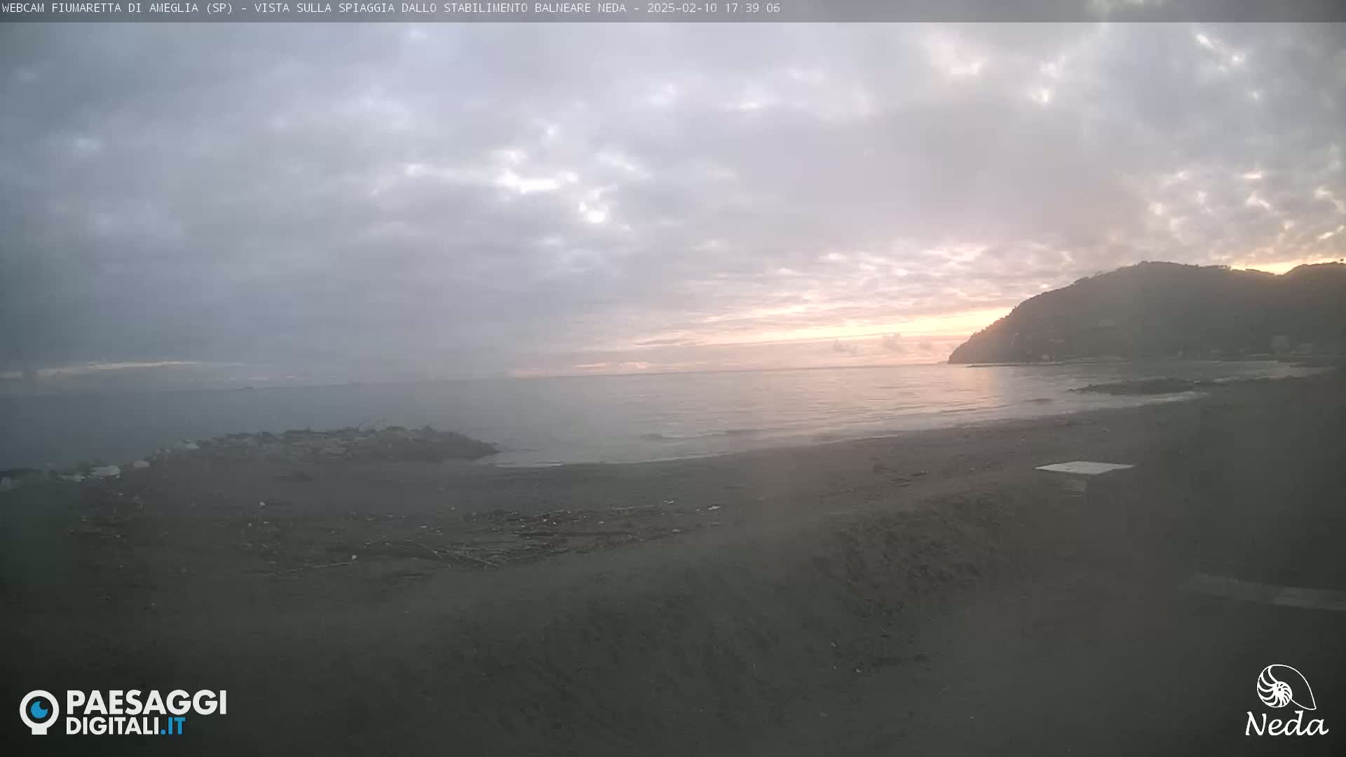 A dark, sandy beach at sunset under a cloudy sky, with a calm sea and a hill in the background.