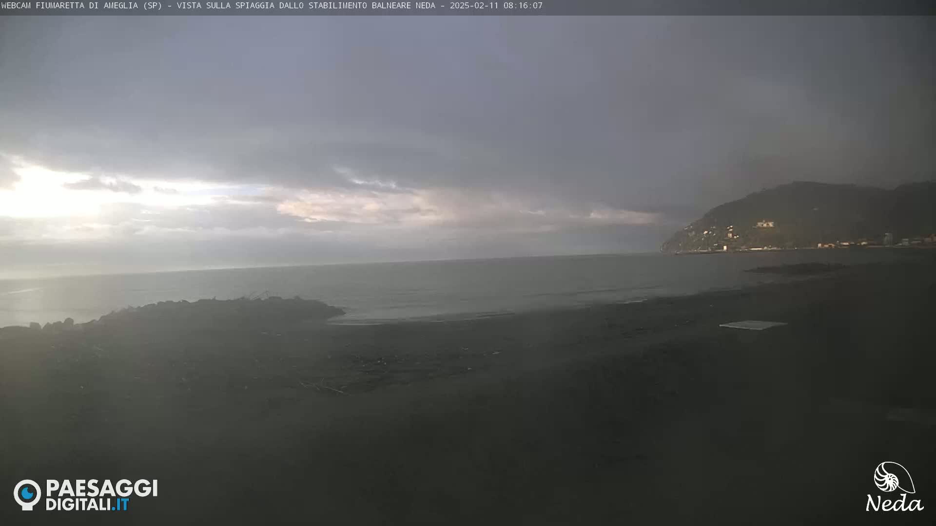 A dark, overcast sky hangs over a calm sea and a dark, sandy beach with a distant hill visible in the background.