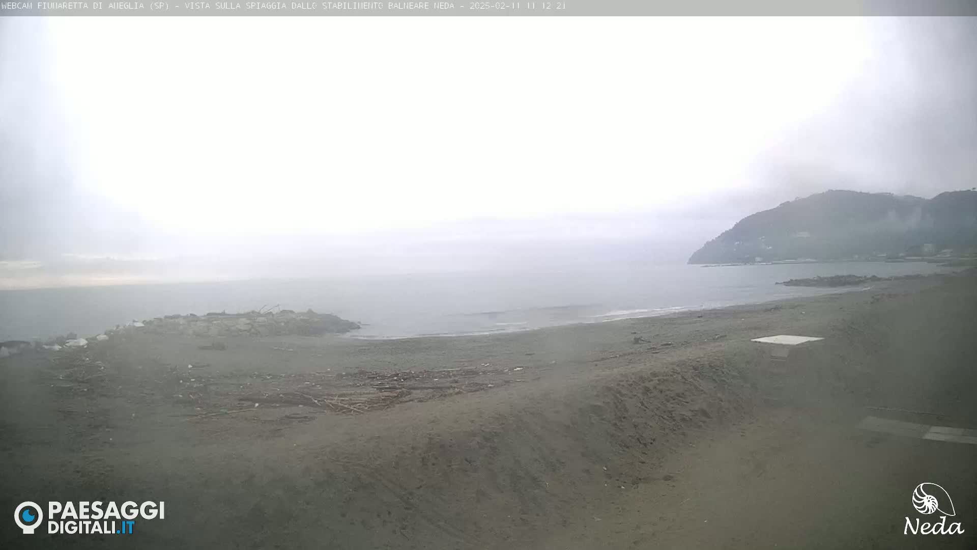 A hazy, overcast day reveals a beach scene with a calm sea, a sandy shore littered with debris, and a hill in the distance.