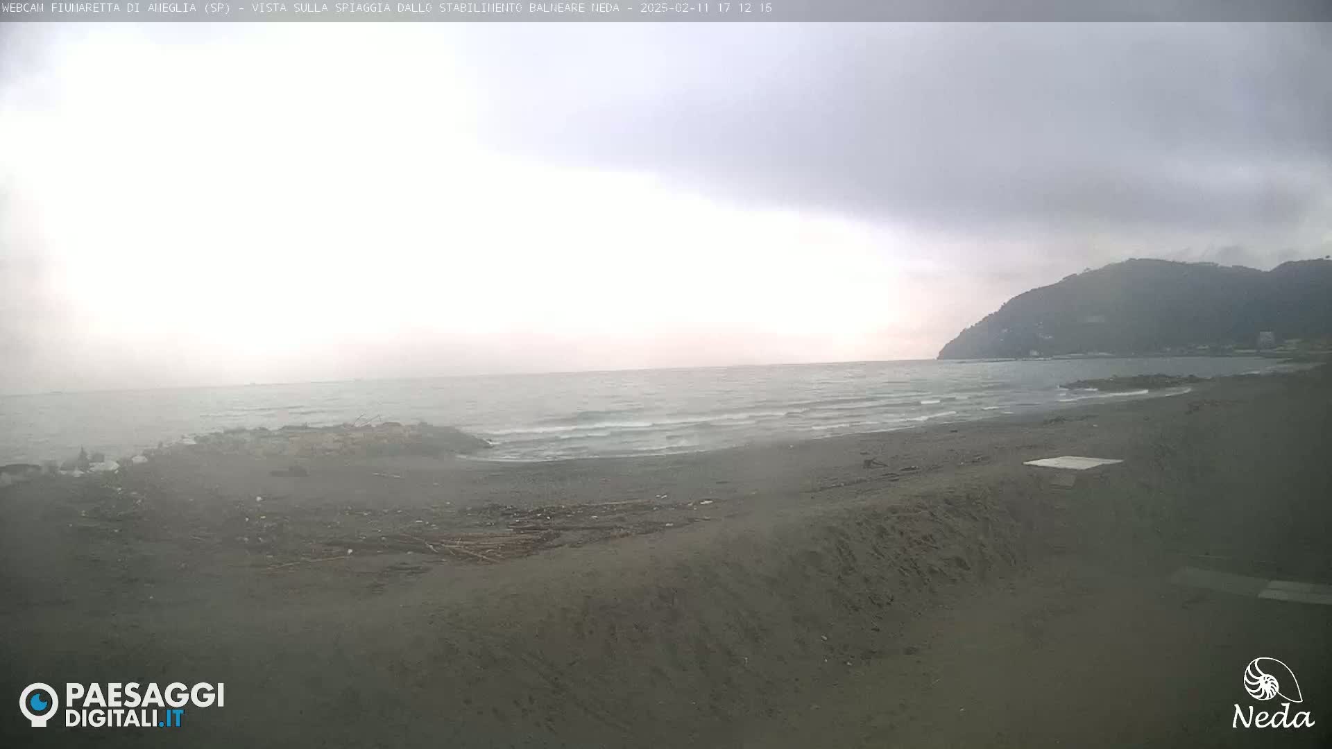 A dark, sandy beach with small waves on a cloudy day, backed by a distant, dark mountain.