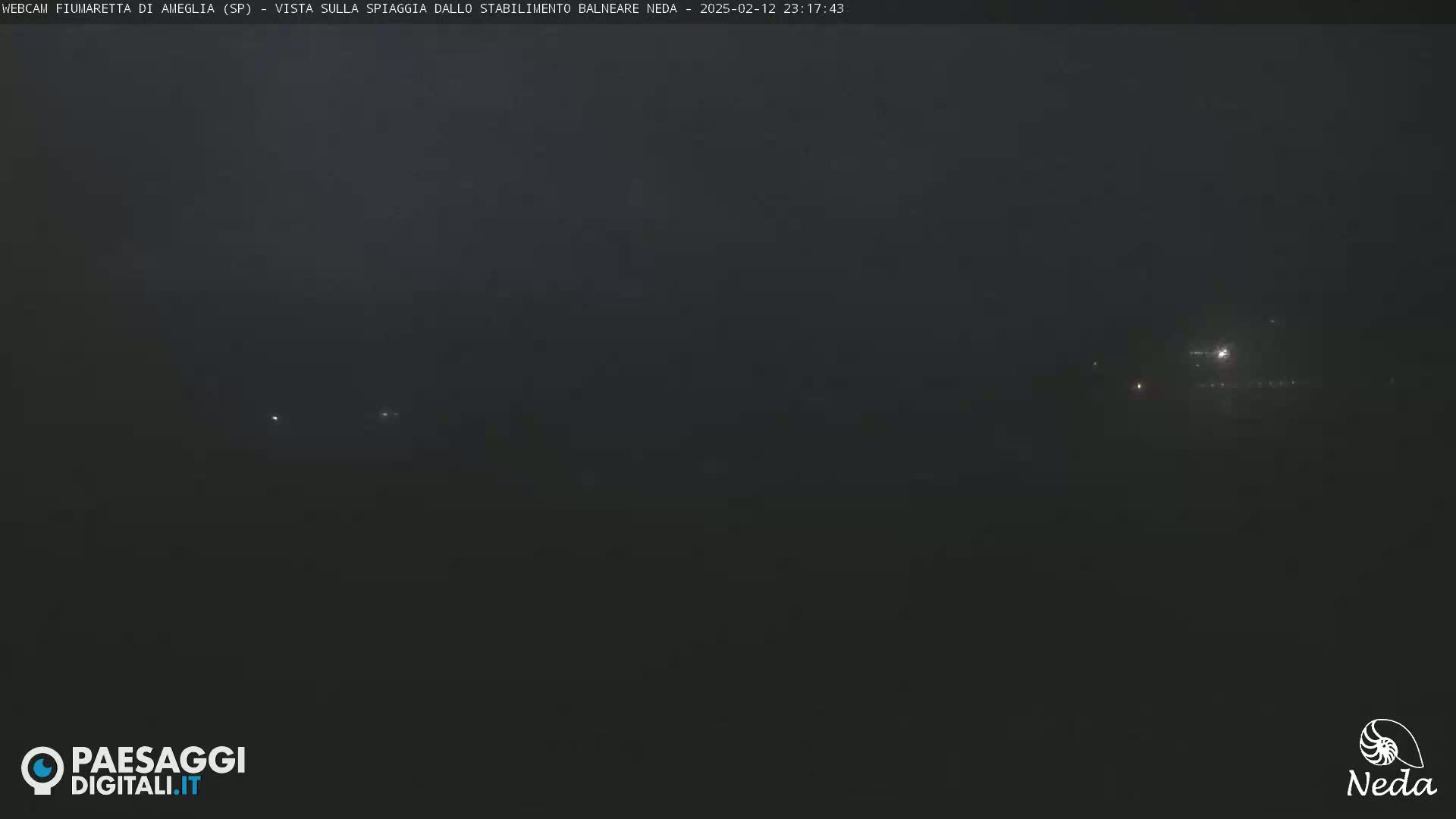 A dark, nighttime view of a beach, possibly obscured by fog or mist, with faint lights visible in the distance.