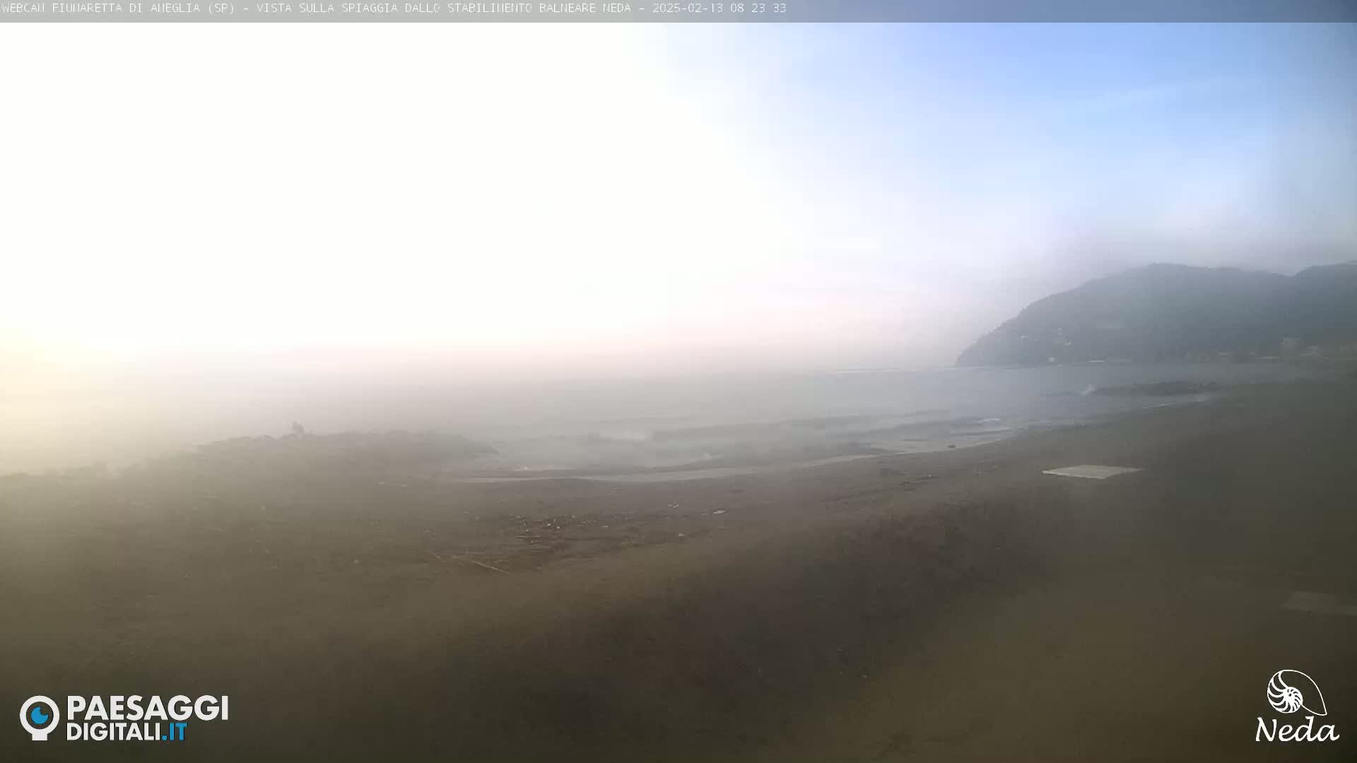A hazy, overcast day at a beach with a dark sandy shore and small waves lapping at the edge of the land, with a distant mountain shrouded in mist.