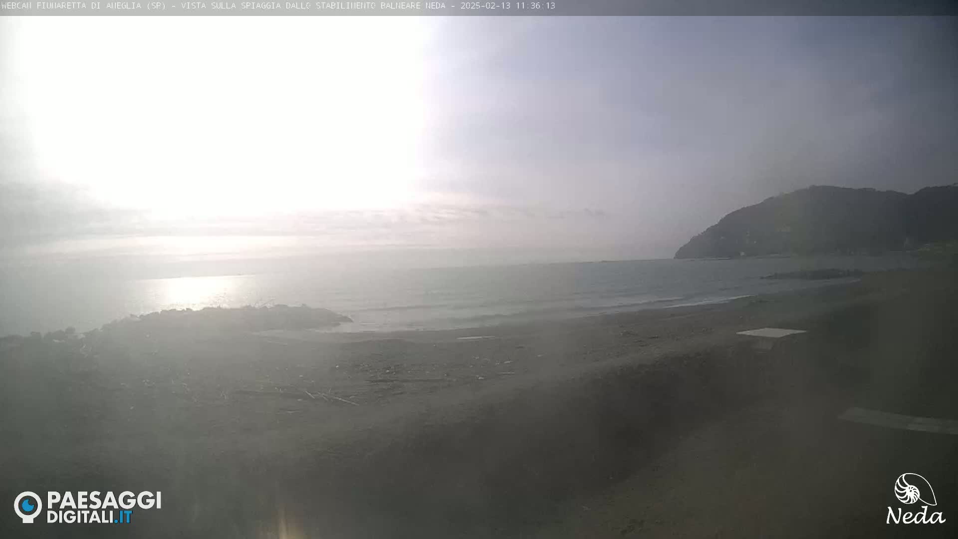 A hazy, overcast day at a beach shows a calm sea with small waves lapping a dark sandy shore, and a distant hill in the background.