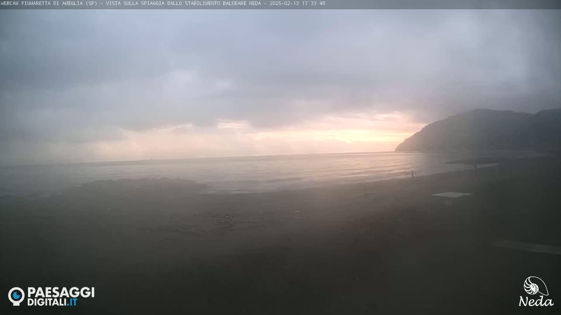 A hazy, overcast beach scene at sunset shows a calm ocean meeting a dark sandy shore with a hill in the background.