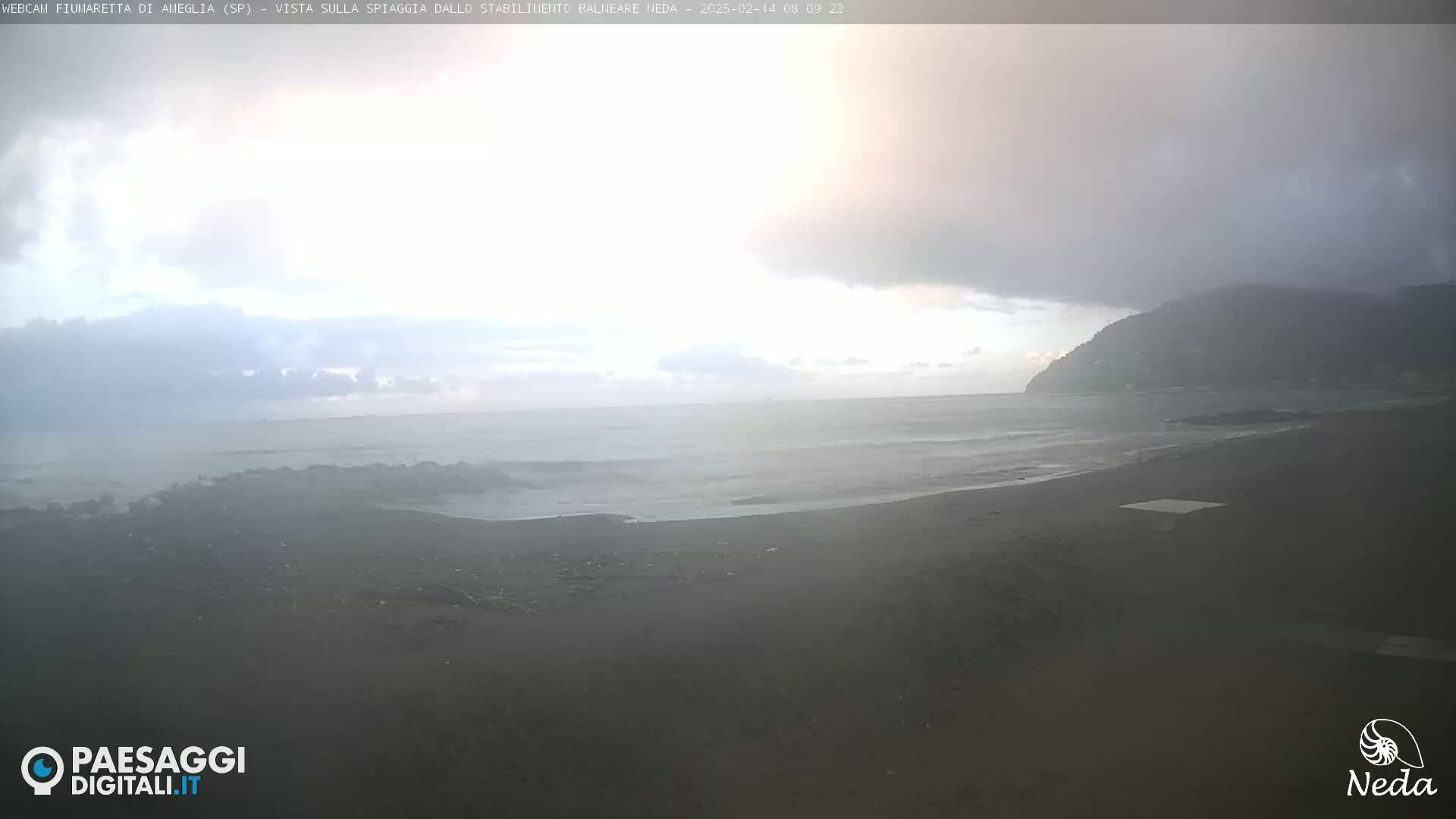 A dark, overcast day shows a beach with dark sand and small waves lapping the shore, with a distant hill shrouded in mist.