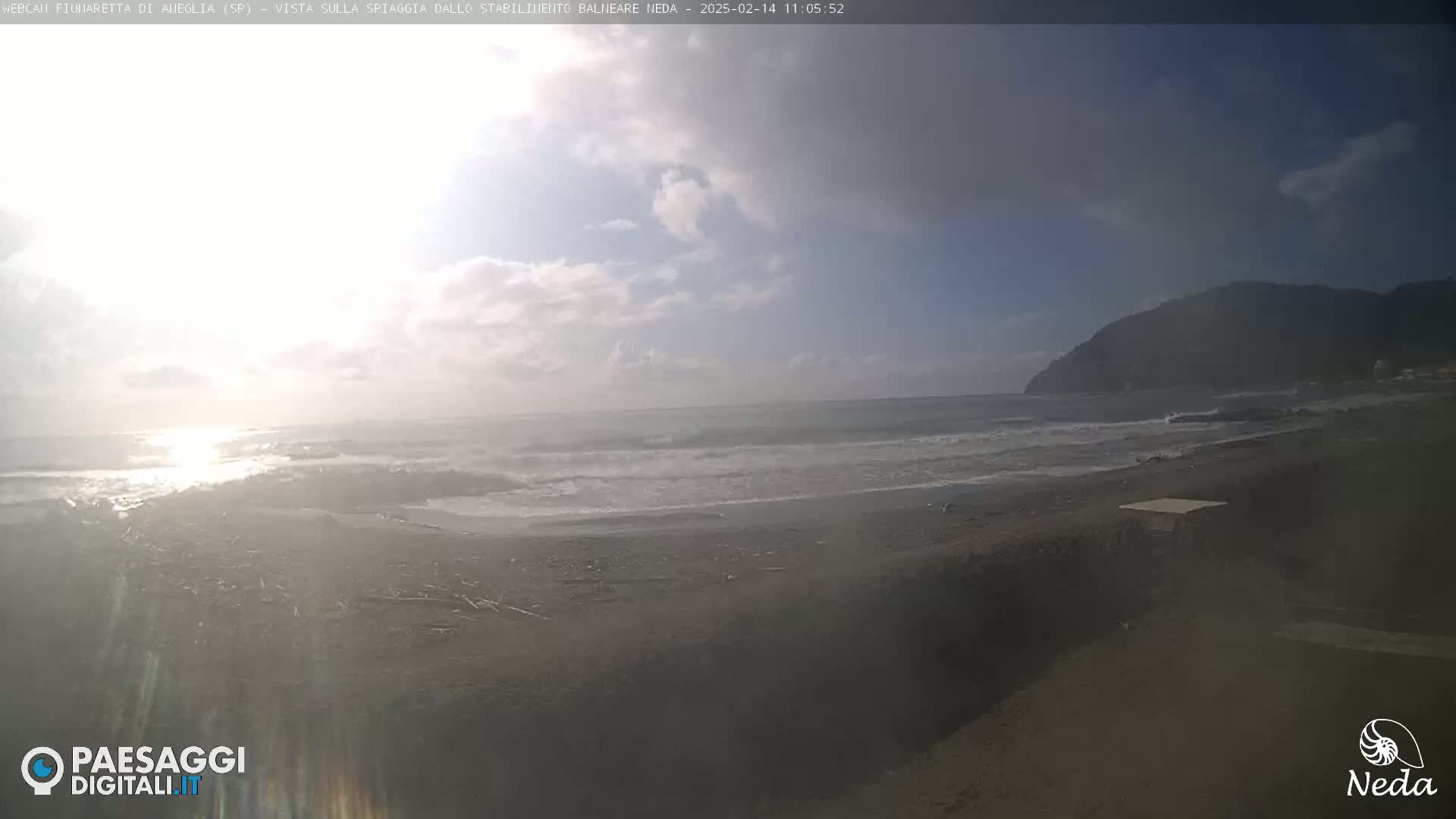 A partly cloudy day shows a beach with moderate waves lapping the shore, backed by a distant mountain.