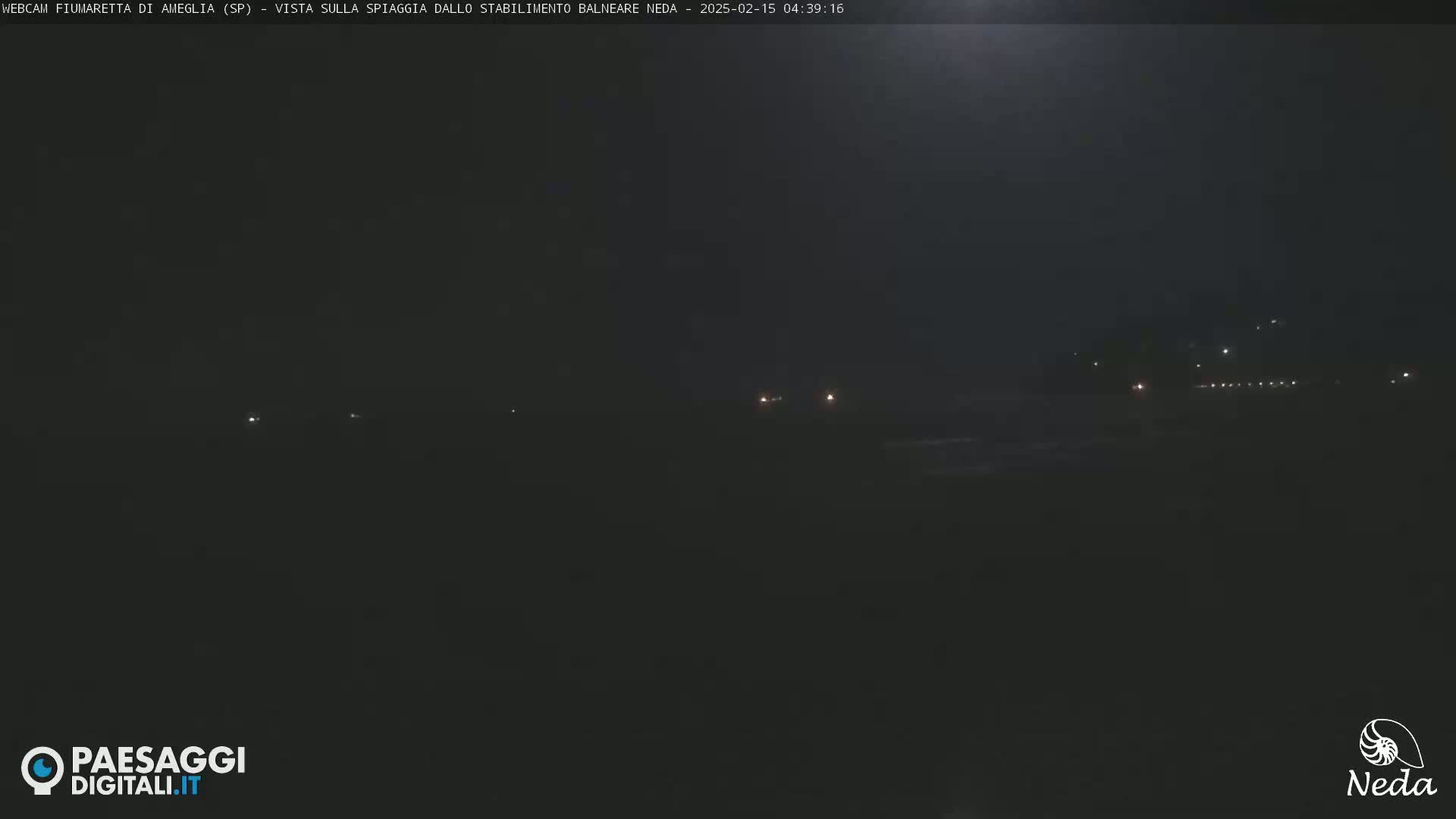A dark, nighttime view of a beach with a few distant lights visible under a mostly cloudy sky.