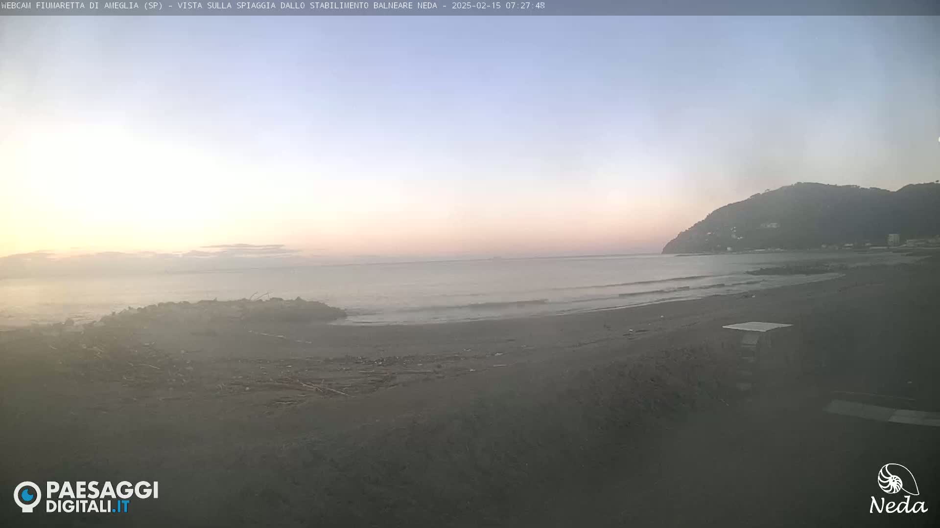 A hazy sunrise illuminates a dark-sand beach and calm ocean waves, with a hill visible in the background.