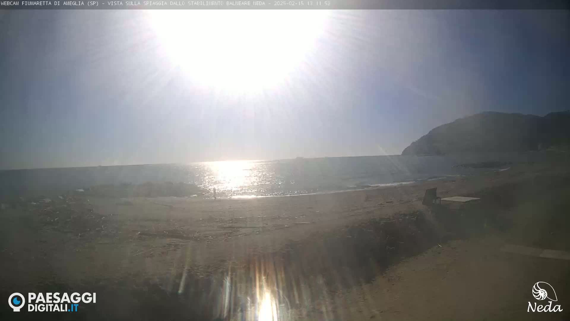 A sunny beach scene shows a calm sea reflecting sunlight, with a sandy shore and a distant hill under a bright sky.
