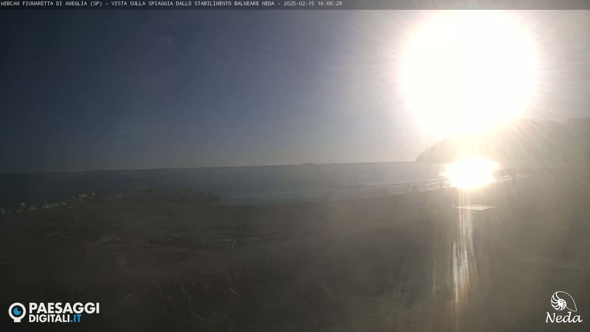 A sunny day at a beach with calm ocean water and a bright sun partially obscured by lens flare.