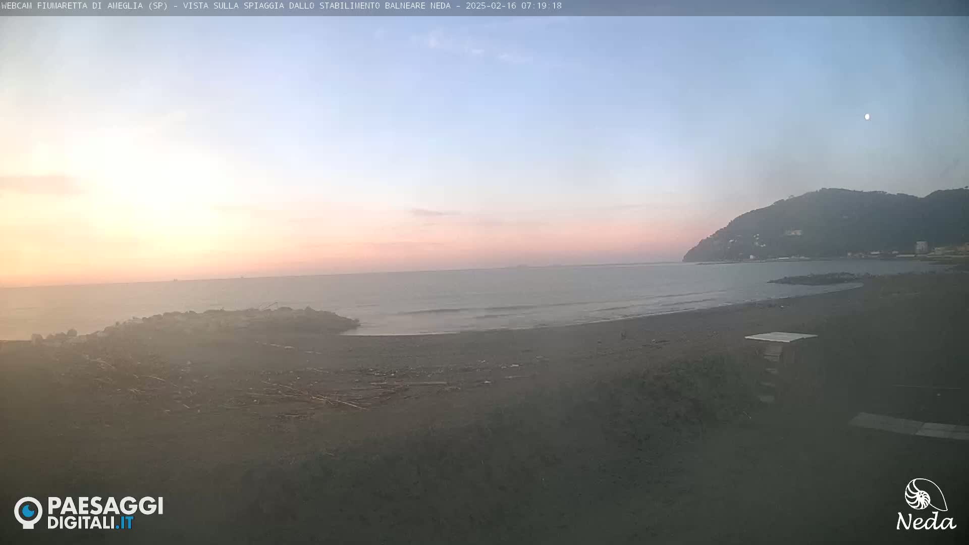 A dark beach with a calm ocean under a pale pink and blue twilight sky, showing a small hill in the distance.
