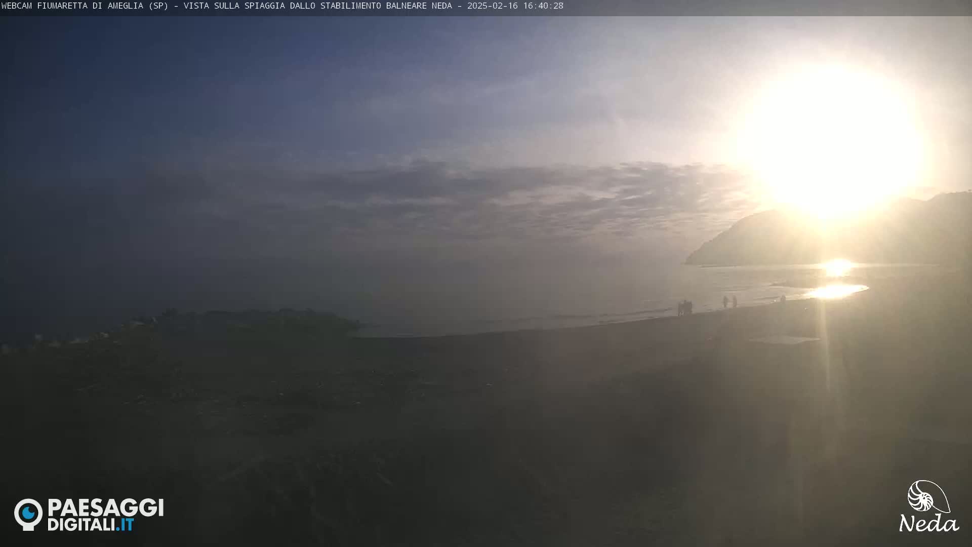 A hazy sunset illuminates a beach scene with a few people visible in the distance.