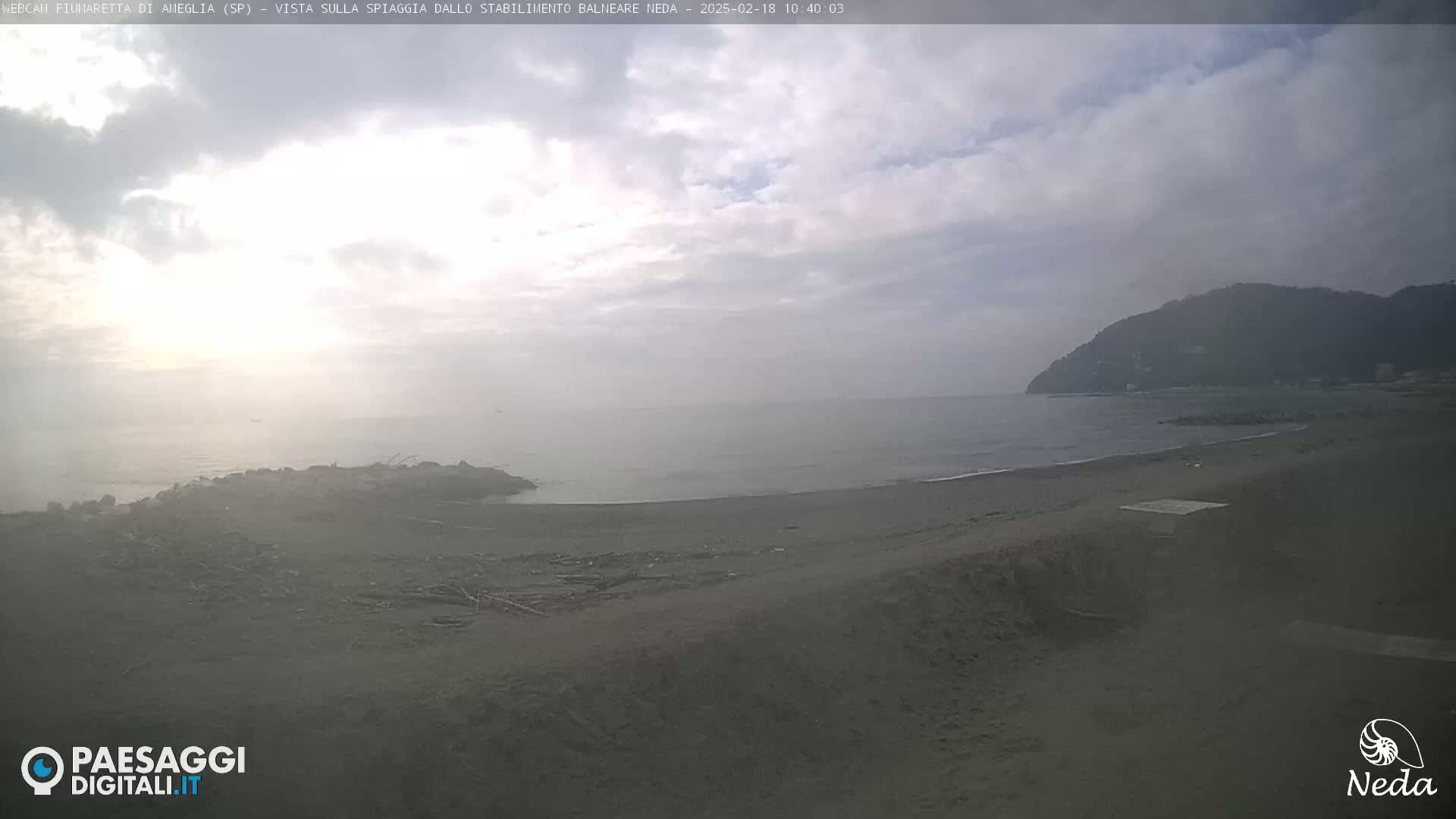 A mostly cloudy day reveals a view of a dark-sand beach and calm sea, with a distant hill visible under a hazy sky.