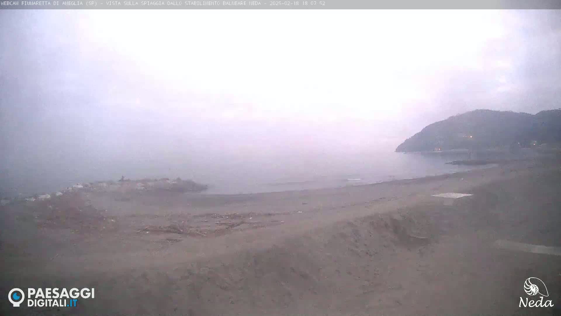 A hazy, overcast day reveals a sandy beach and calm sea with a distant hill in the background.