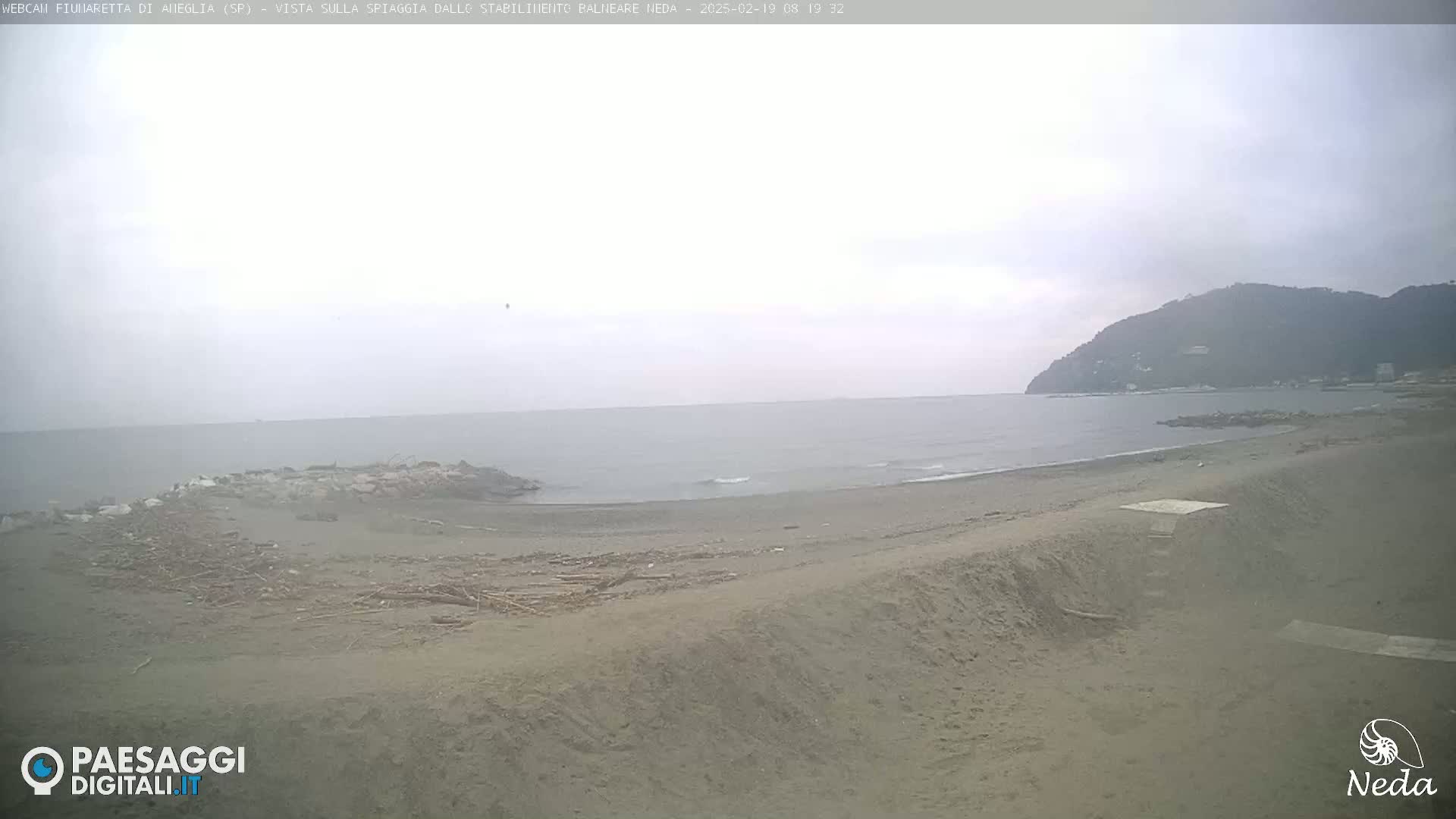 A sandy beach with debris and a calm sea under an overcast sky is visible, with a hill in the background.