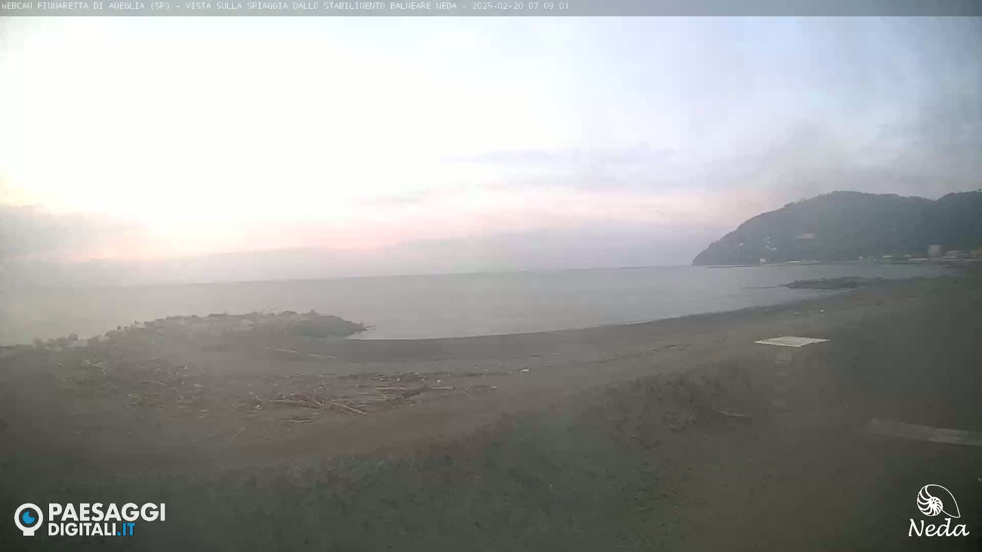 A hazy sunrise over a dark-sand beach and calm sea, with a distant hill visible under a pale sky.