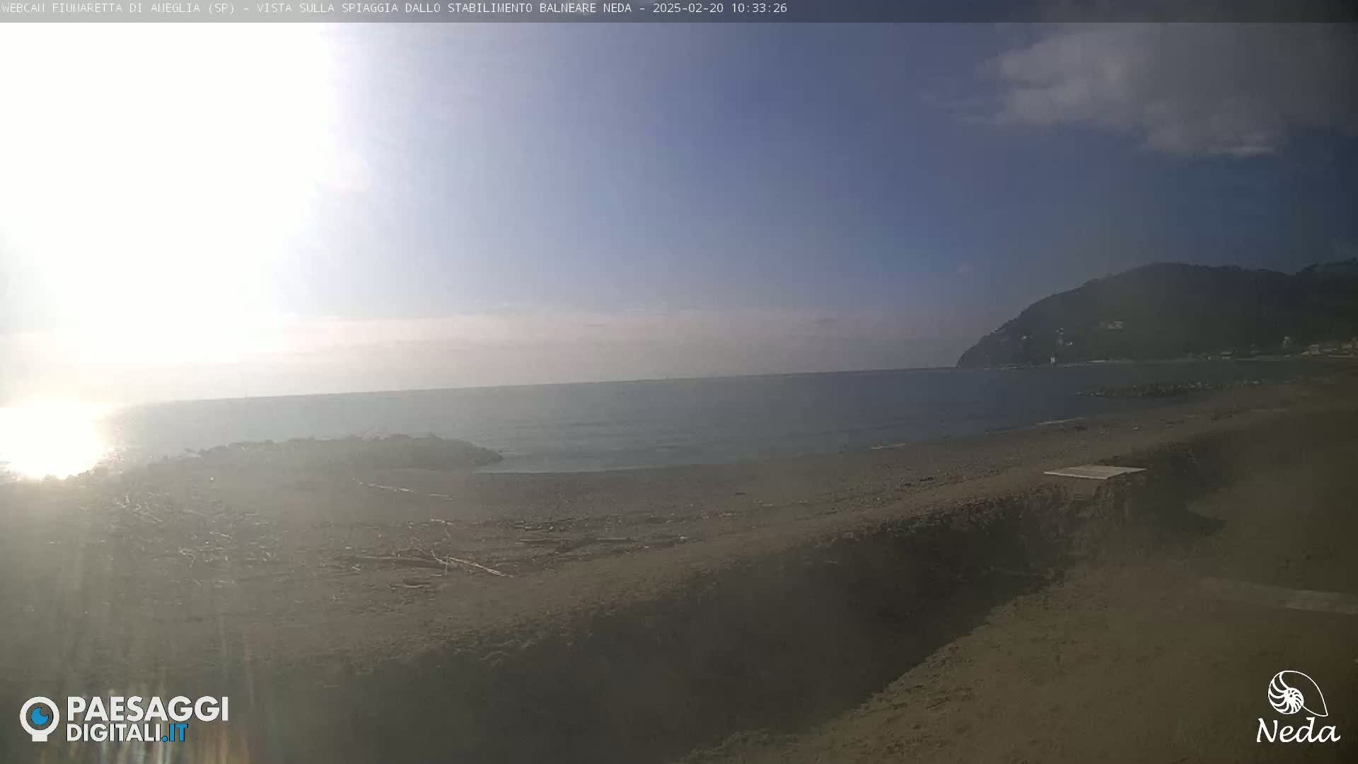 A mostly sunny day at a sandy beach with calm water and a distant mountain in the background.