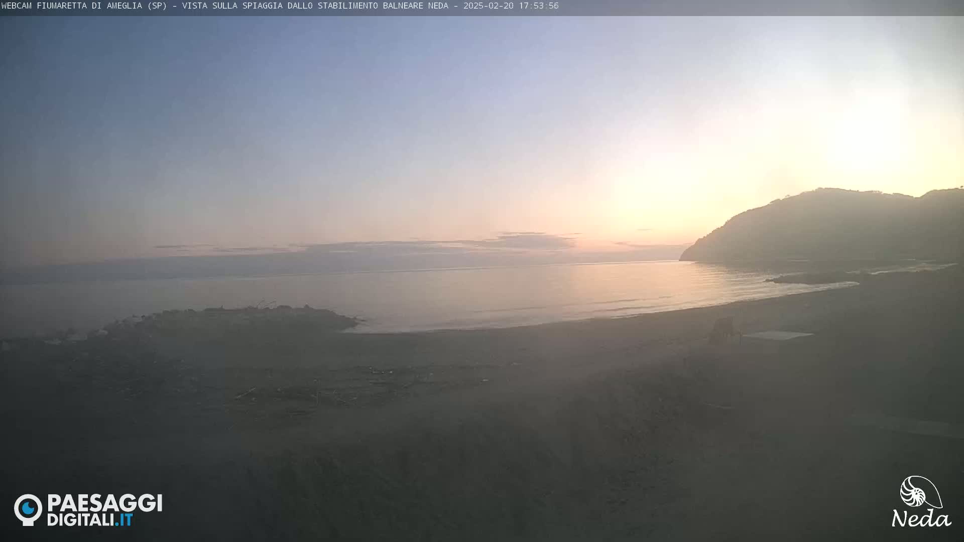 A hazy sunset illuminates a calm ocean beach with a hill in the background.
