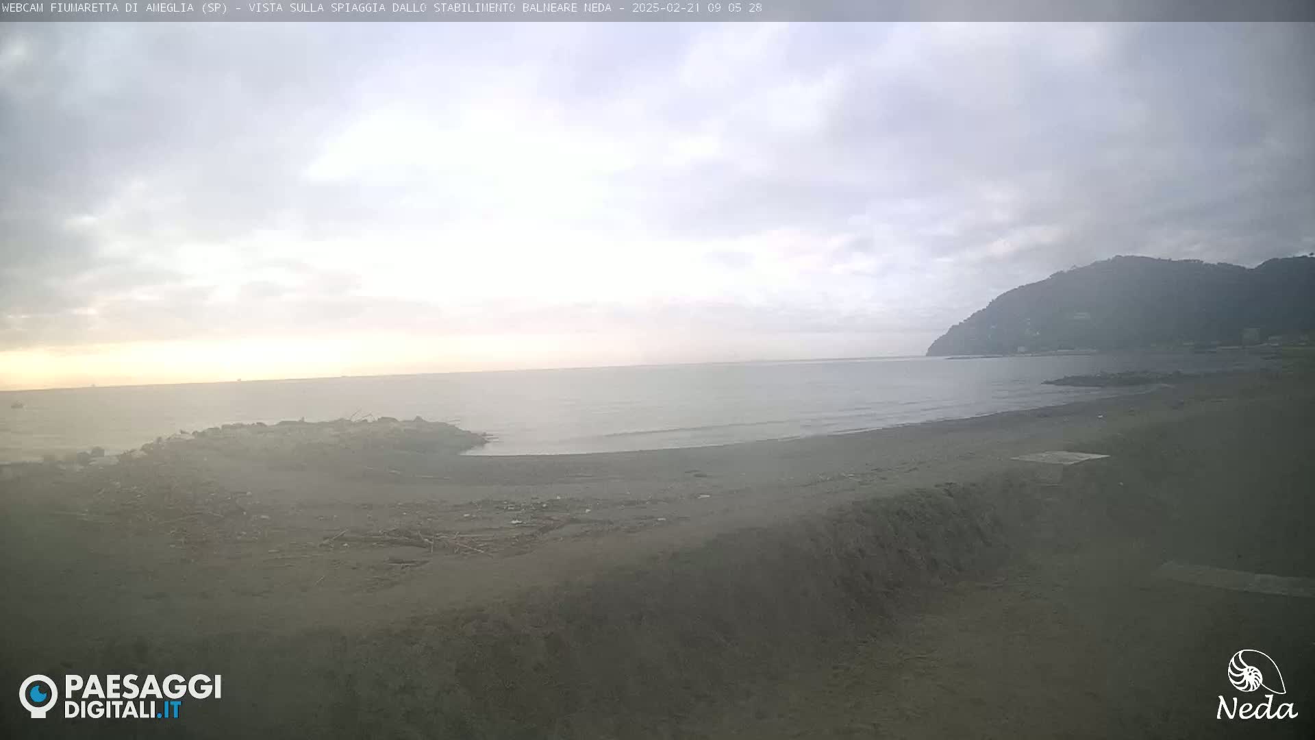 A dark, overcast day shows a view of a mostly empty beach and calm ocean with a hill in the distance.