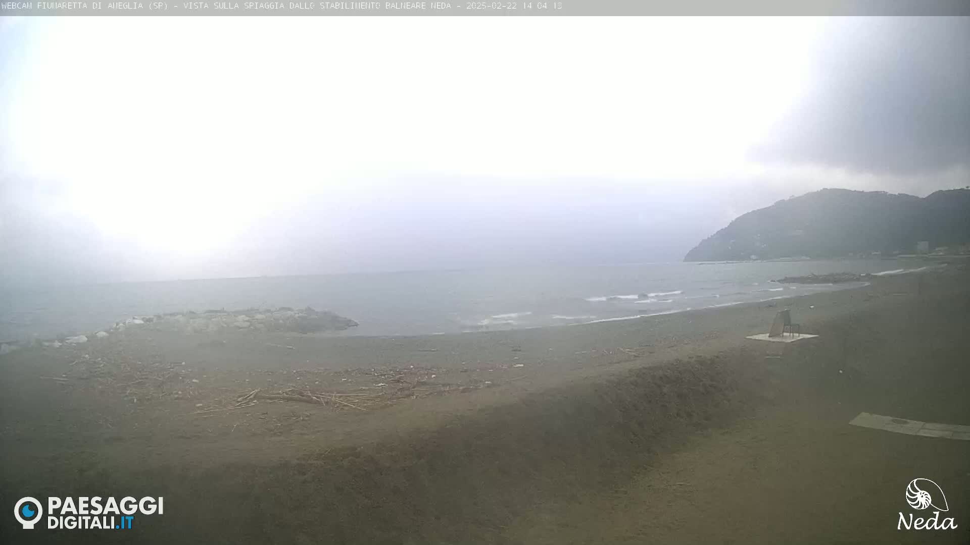 A hazy, overcast day shows a dark-sand beach with small waves lapping the shore, a lone chair on the sand, and a hill in the distance.