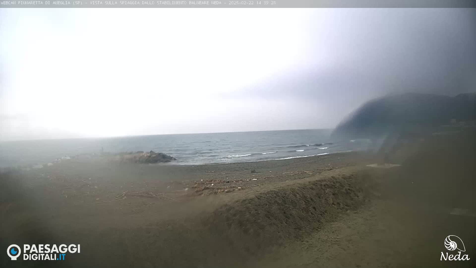 A hazy, overcast day at a beach with a dark, sandy shore and calm waves.
