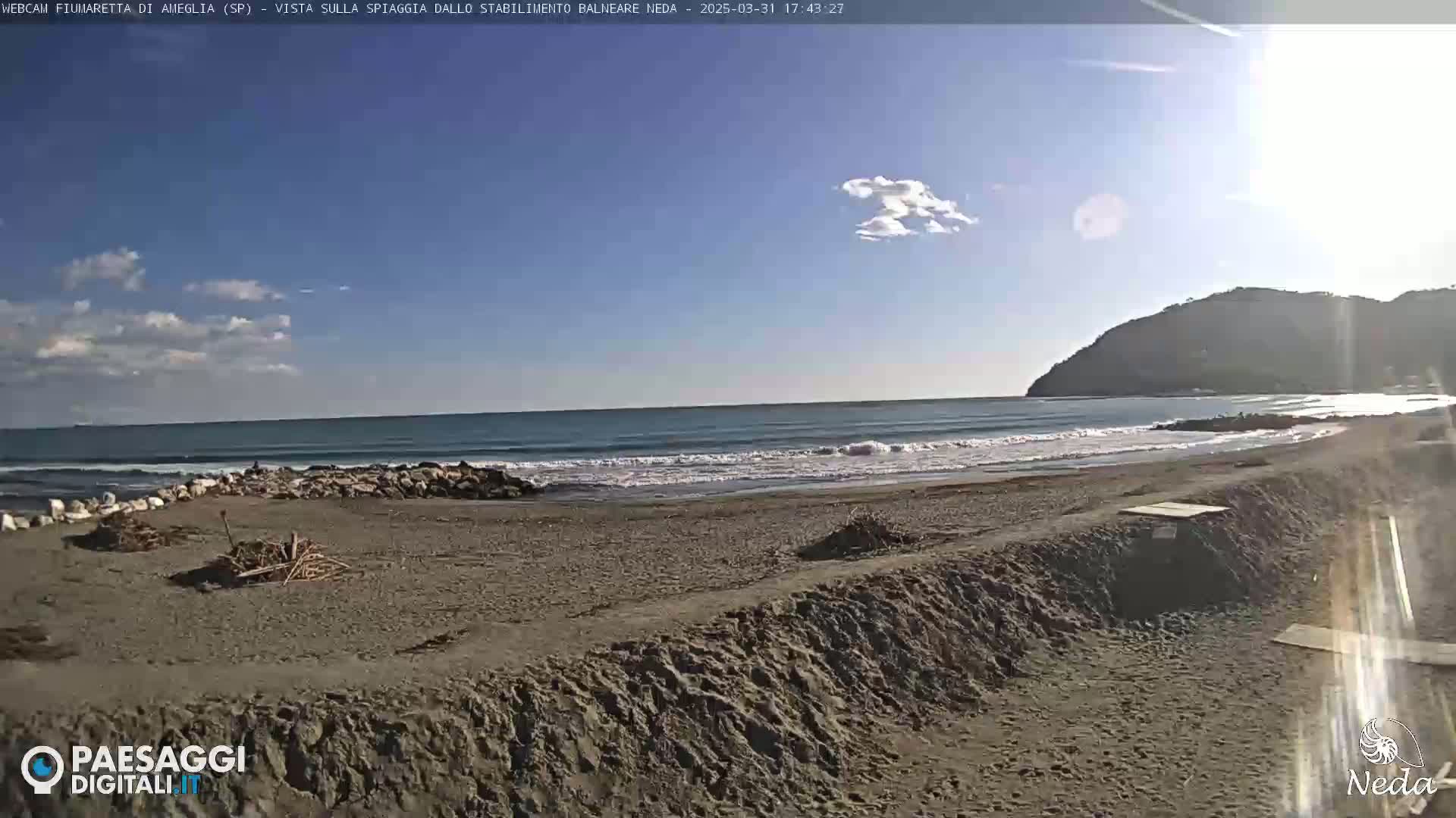 A sandy beach with small waves rolls onto the shore under a mostly clear, sunny sky, with a hill visible in the background.