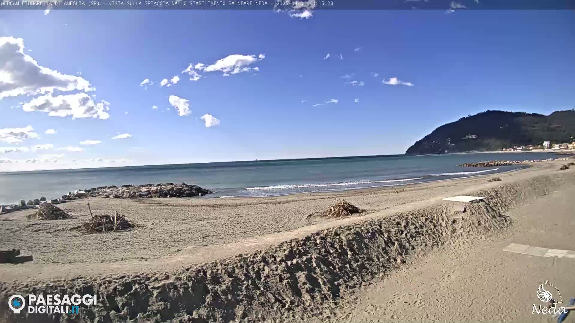 A sandy beach with calm ocean waves under a mostly sunny sky, backed by a hill.