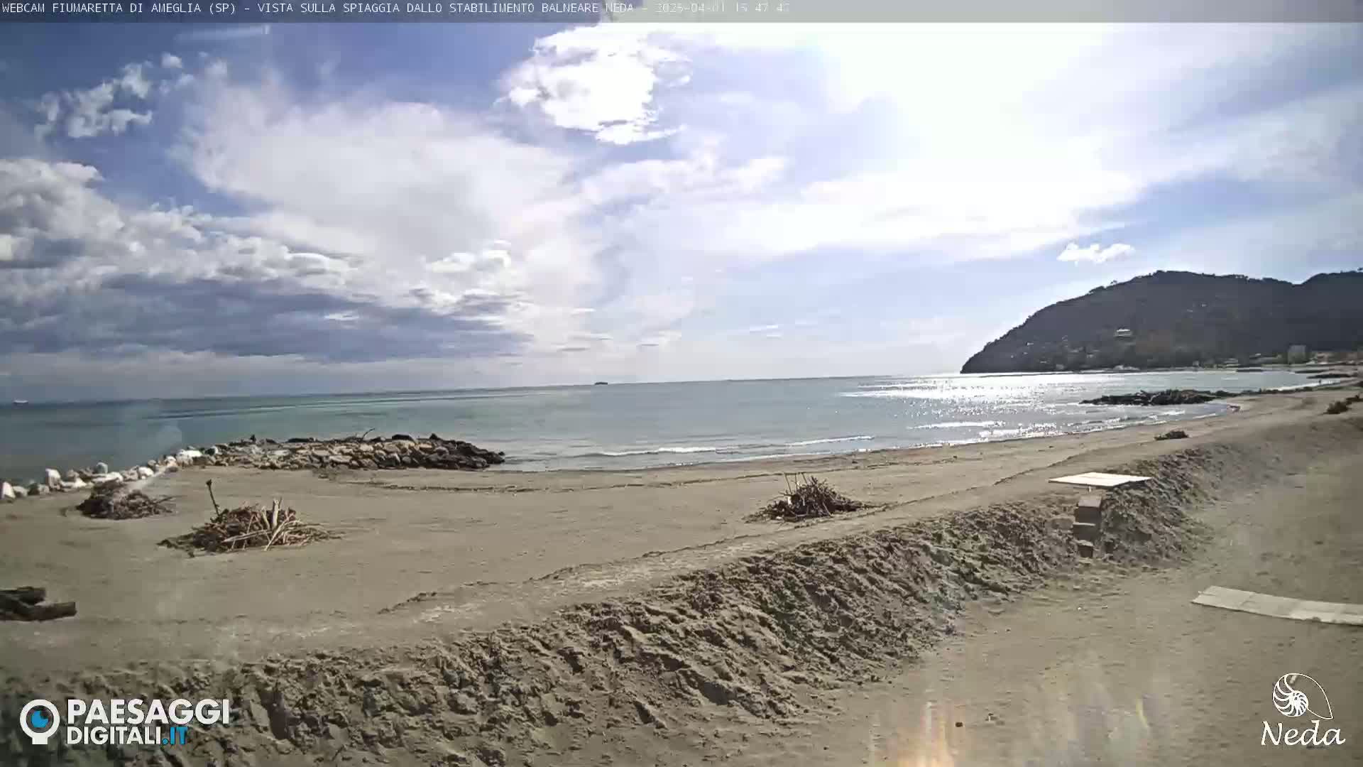 A partly cloudy day shows a sandy beach with small piles of driftwood, a calm sea, and a distant hill.