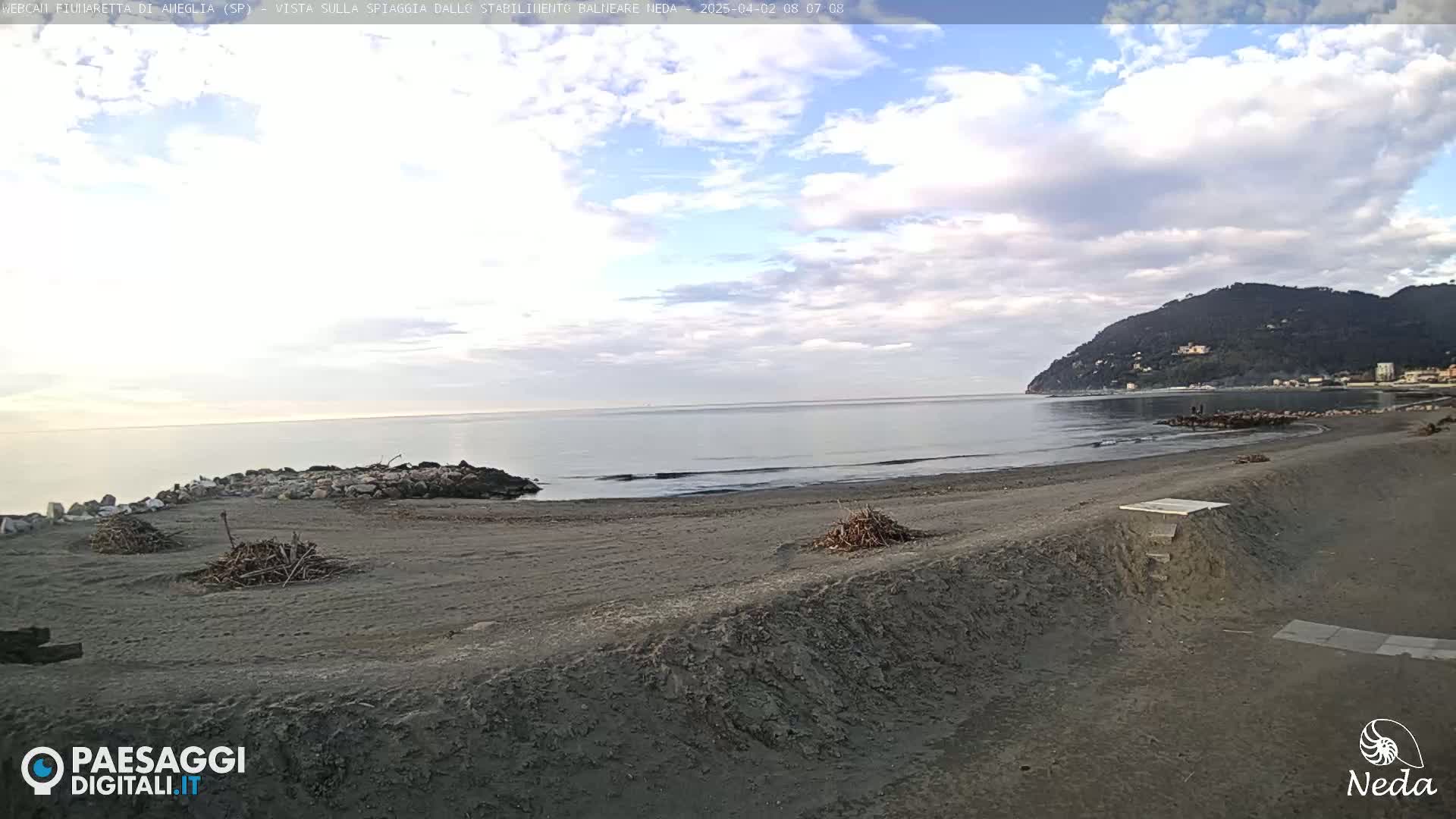 A mostly cloudy day shows a sandy beach and calm ocean with a distant mountain range under a partly cloudy sky.