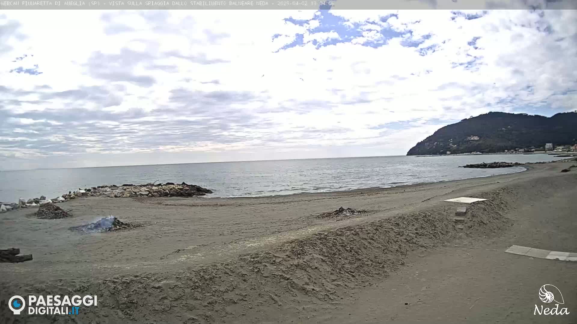 A sandy beach with a calm sea and a hill in the background under a partly cloudy sky.