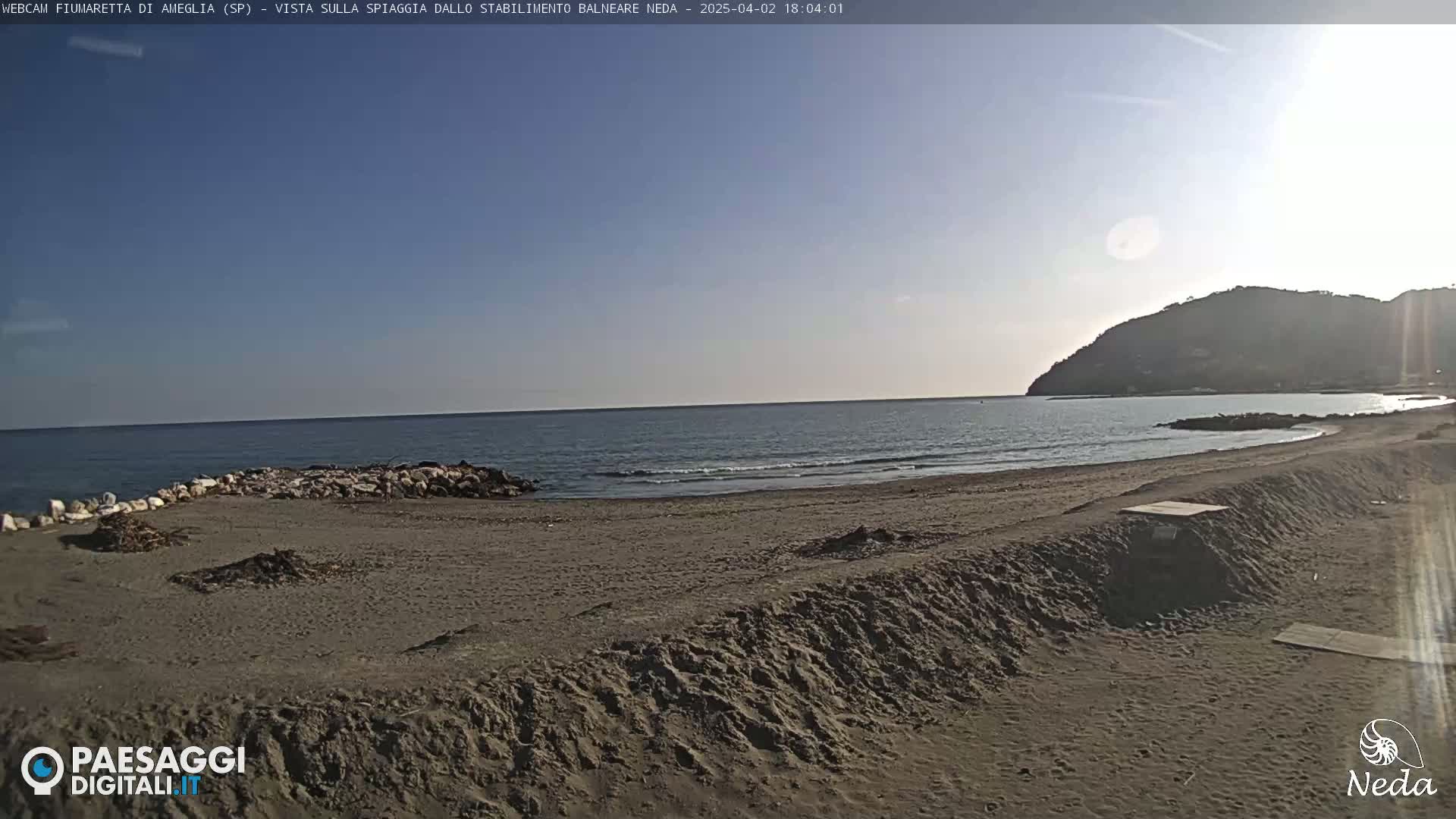 A sandy beach stretches along a calm ocean under a clear, sunny sky, with a distant hill in view.