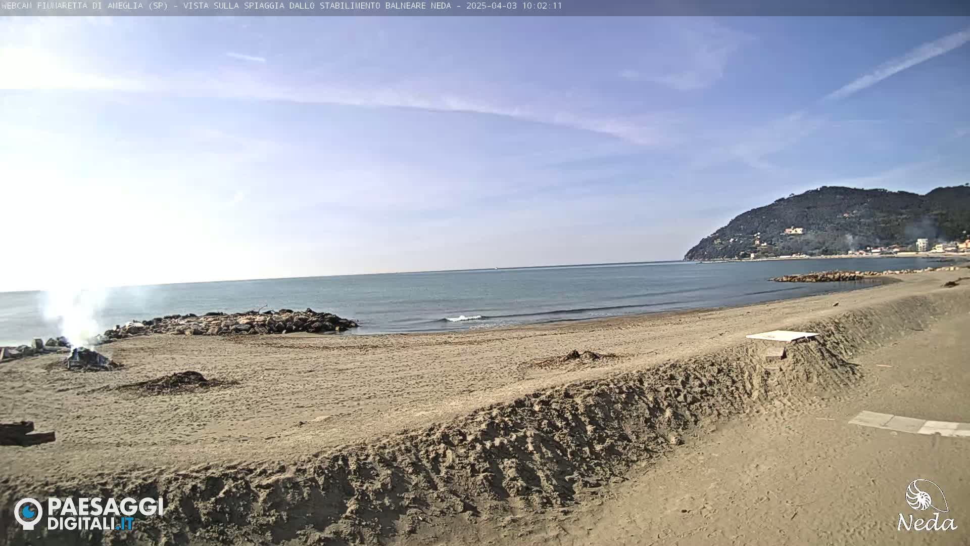 A sandy beach with a calm sea under a mostly clear sky shows a small fire burning near some rocks on the shore, with hills visible in the distance.