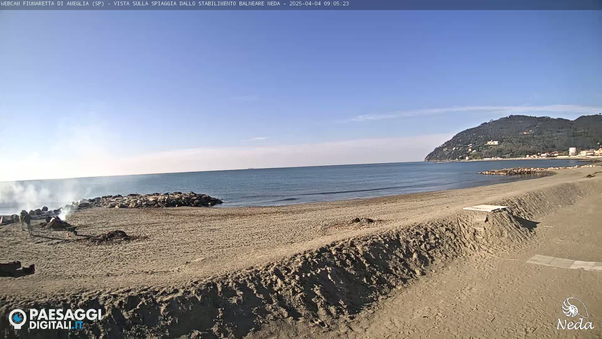A sandy beach with a calm sea under a clear blue sky, a small fire burning near the water, and a hill visible in the distance.