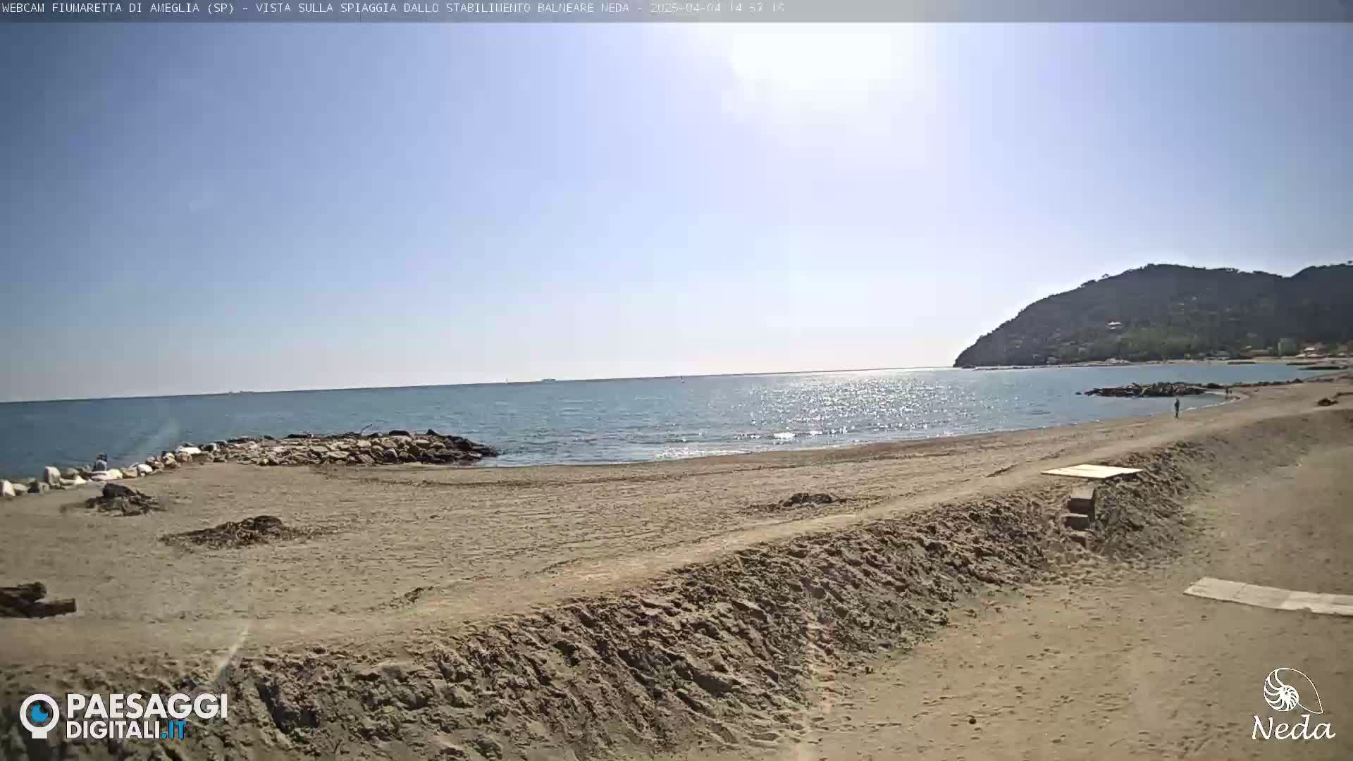 A sandy beach stretches towards a calm, sunlit sea under a clear sky, backed by a distant, verdant hill.