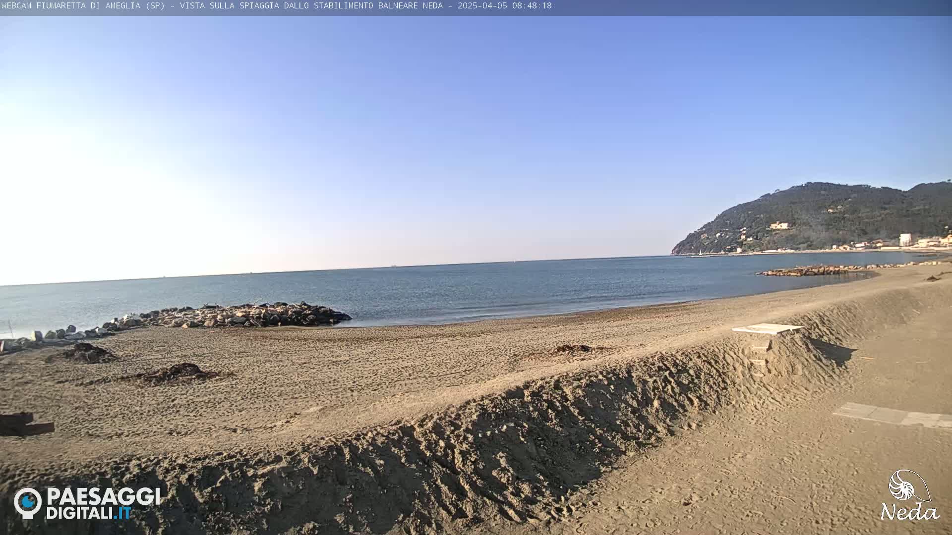 A sandy beach stretches towards a calm sea under a clear blue sky, with a hill visible in the distance.