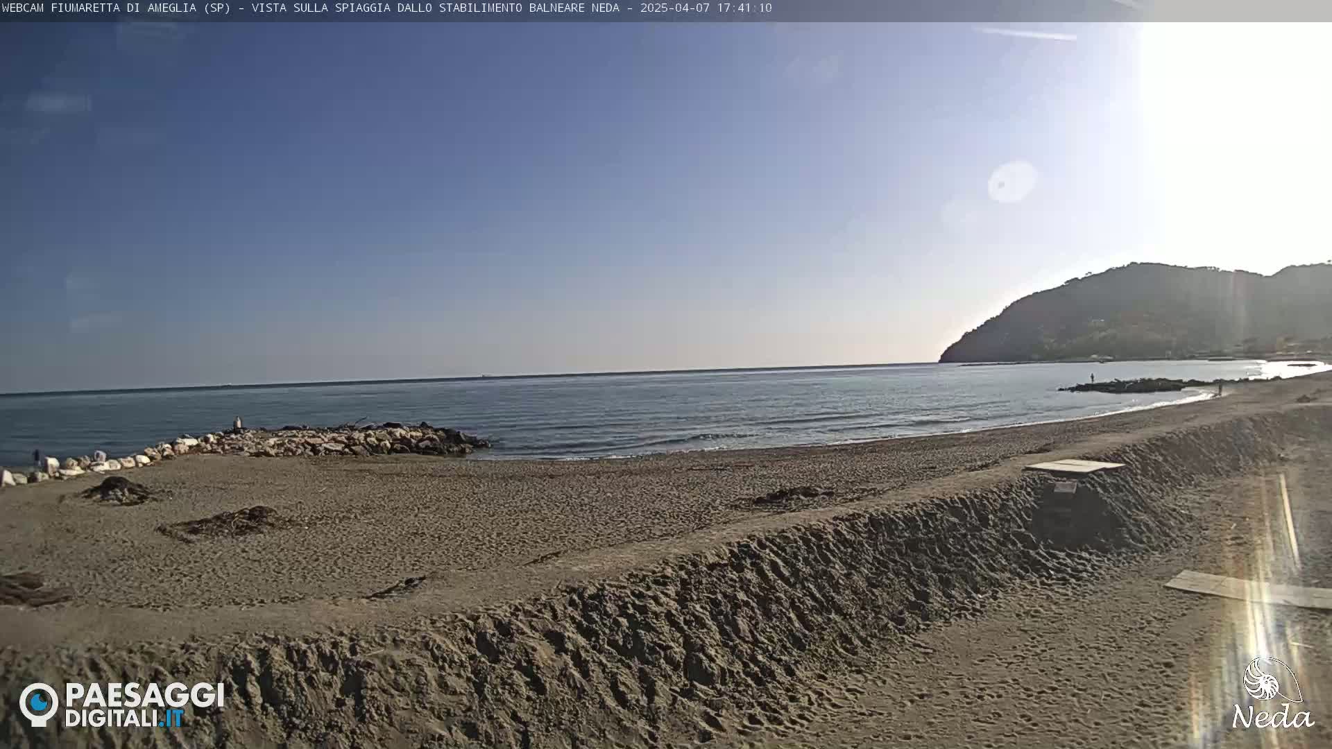 A sandy beach with a calm sea under a clear blue sky, backed by a low hill.