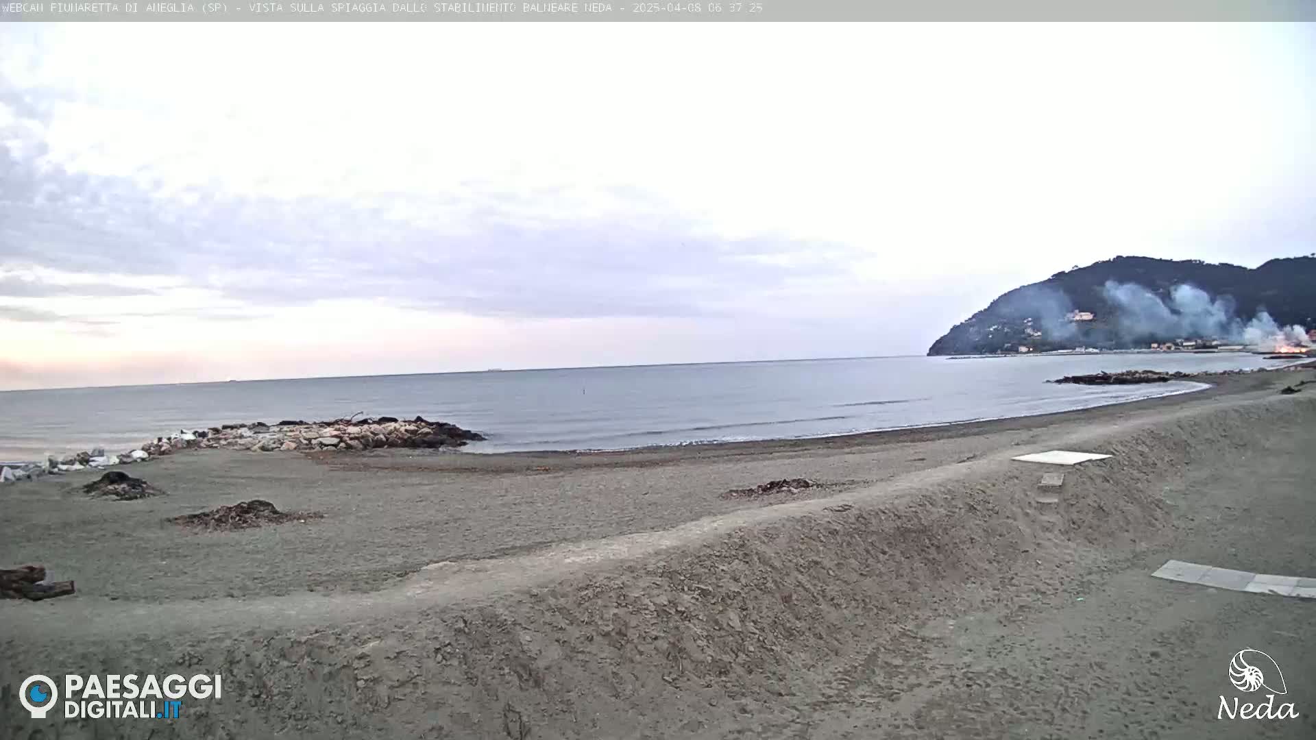 A sandy beach stretches along a calm sea under an overcast sky, with a hill in the distance showing smoke.
