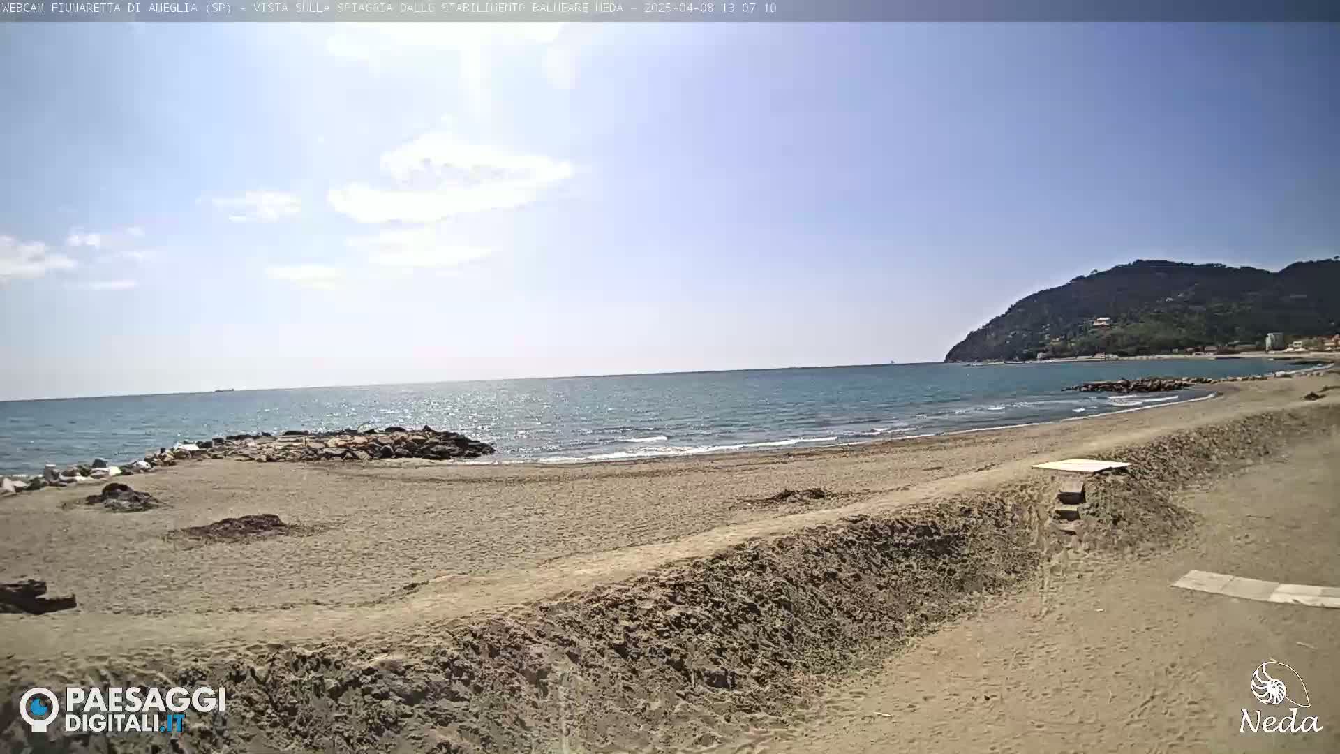 A sandy beach stretches along a calm sea under a mostly clear sky, with a hill visible in the distance.