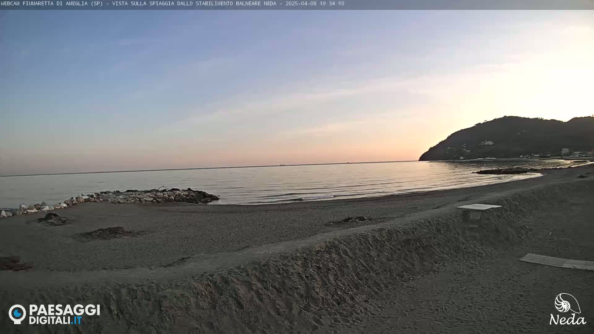 A tranquil beach scene at sunset shows a calm sea lapping a dark-sand shore, with a hill visible in the distance.