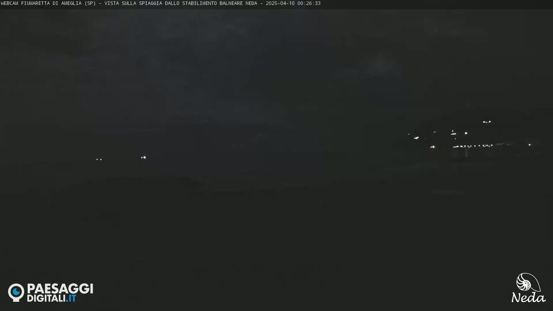 A dark nighttime scene shows a distant line of lights on a hill above a dark beach.