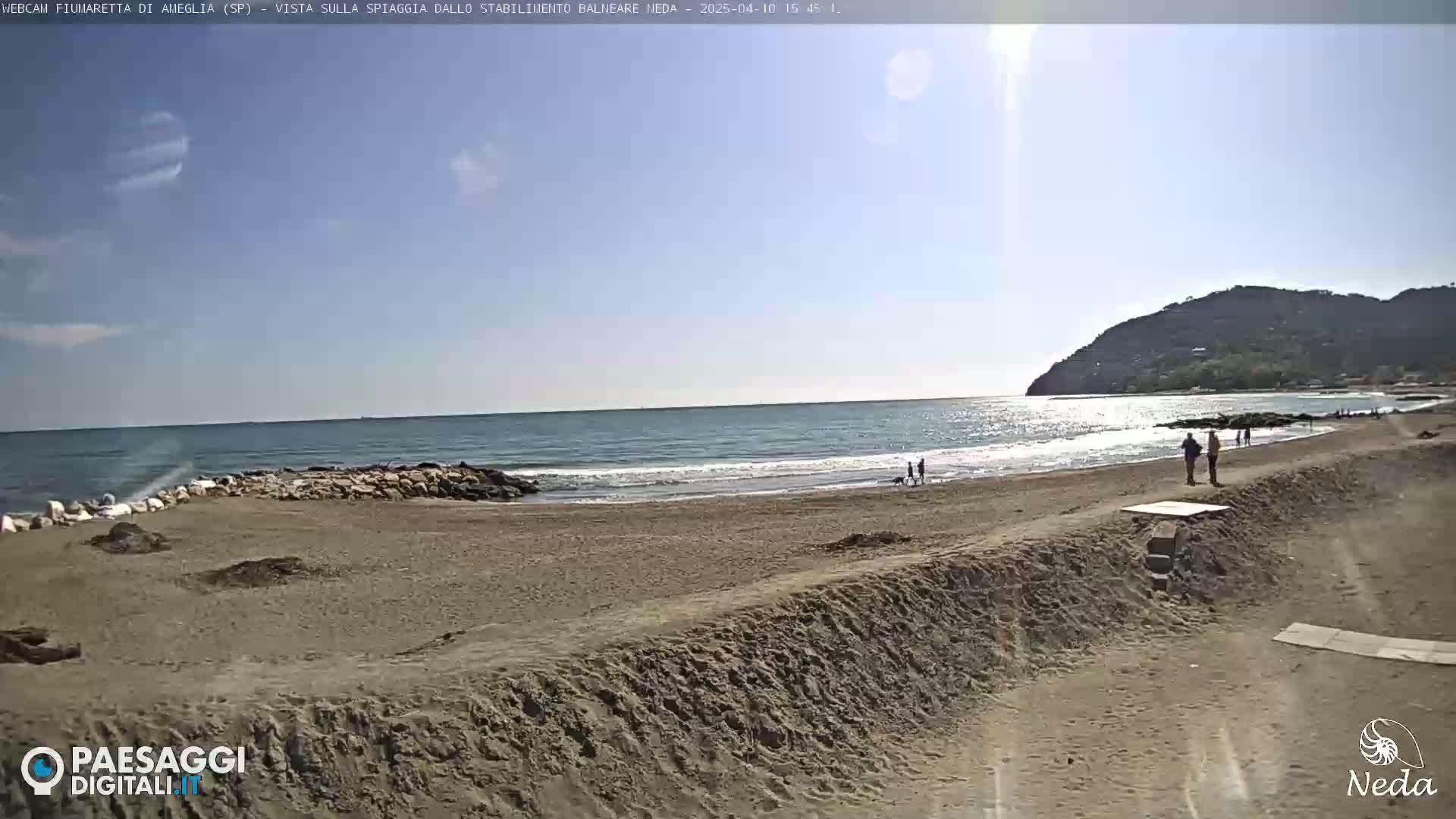 A sandy beach with a few people on it stretches out to a calm sea under a mostly sunny sky, with a hill visible in the background.