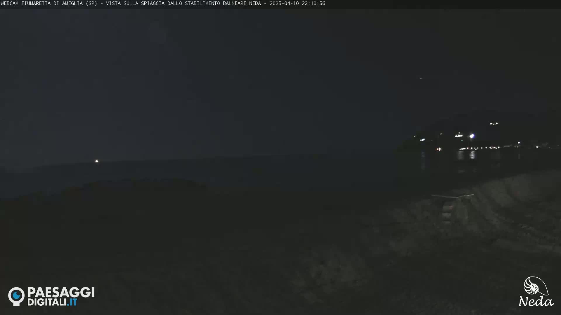 A nighttime, dark view of a beach with distant city lights visible across a body of water.