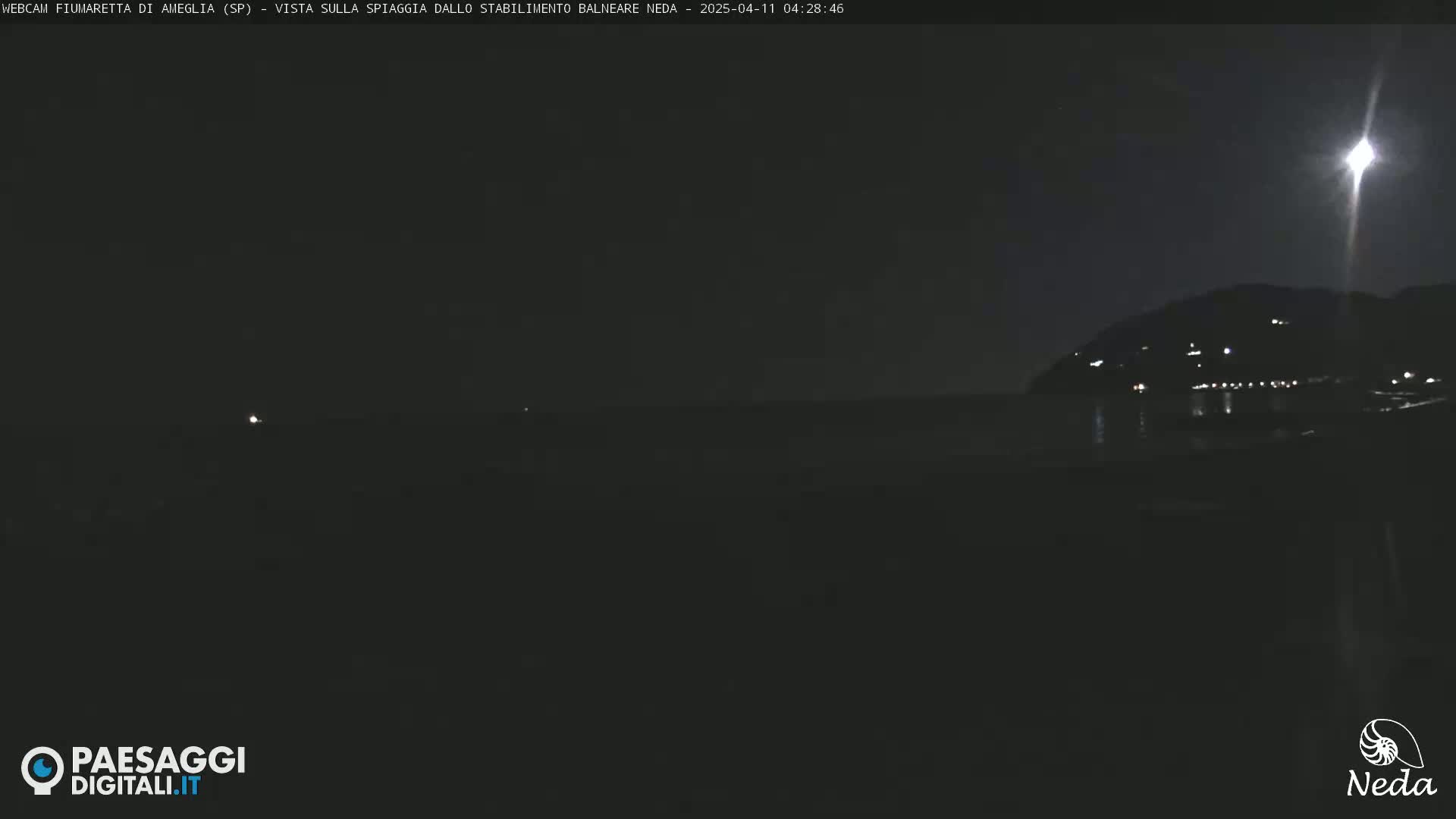 A nighttime view of a dark beach and calm ocean under a bright moon, with a distant hillside showing faint lights.