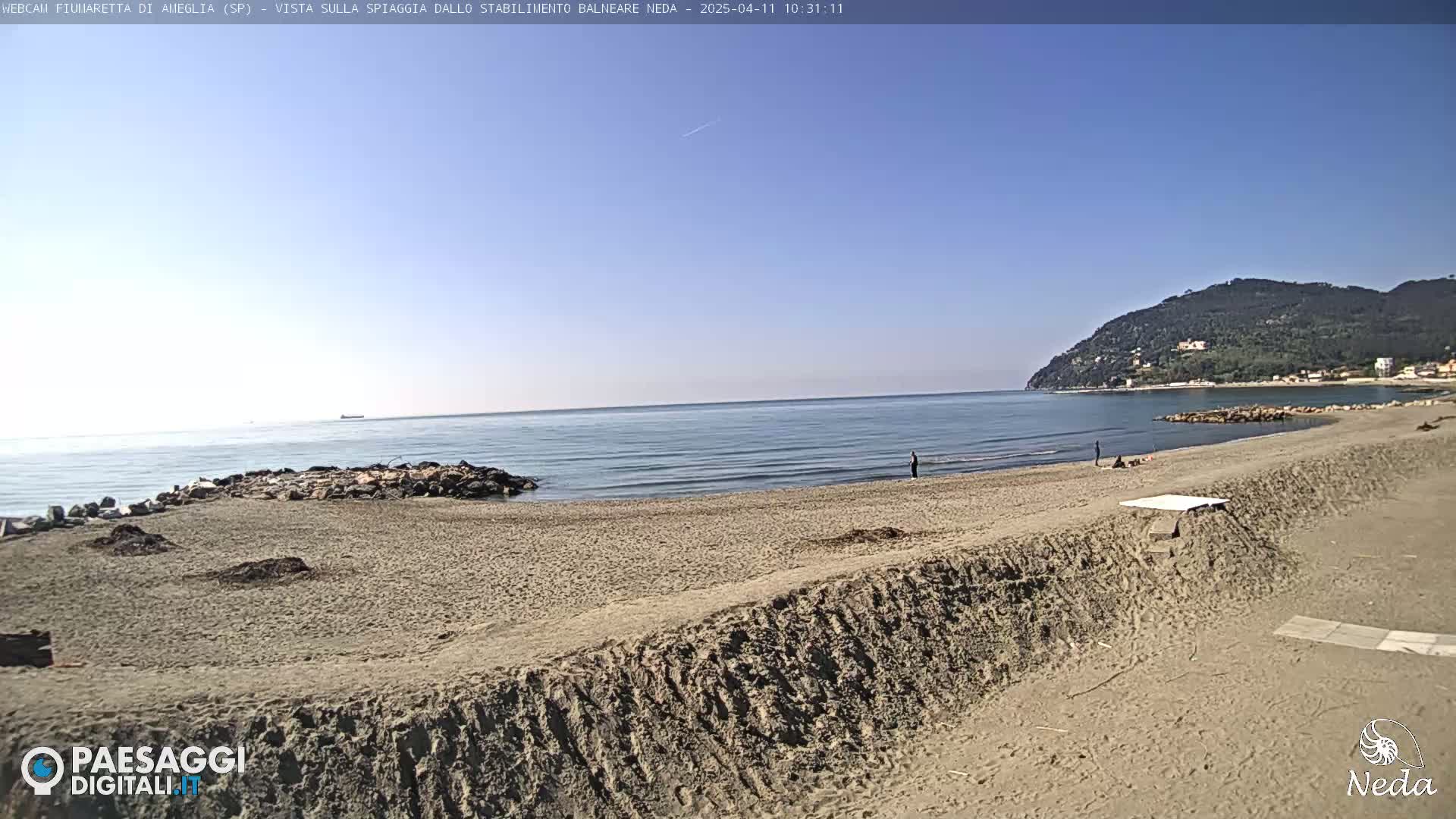 A sandy beach stretches along a calm ocean under a clear blue sky, with a hill visible in the distance.