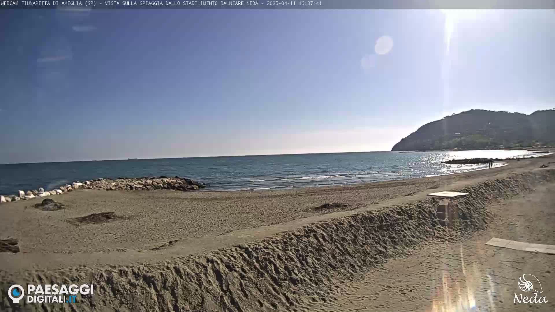 A sandy beach stretches along a calm sea under a bright, sunny sky, with a hill visible in the distance.