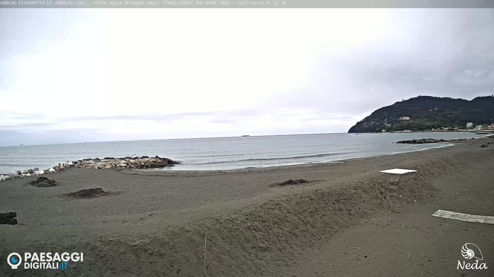 A dark-sand beach stretches towards a calm, gray sea under an overcast sky, with a distant hill visible on the horizon.