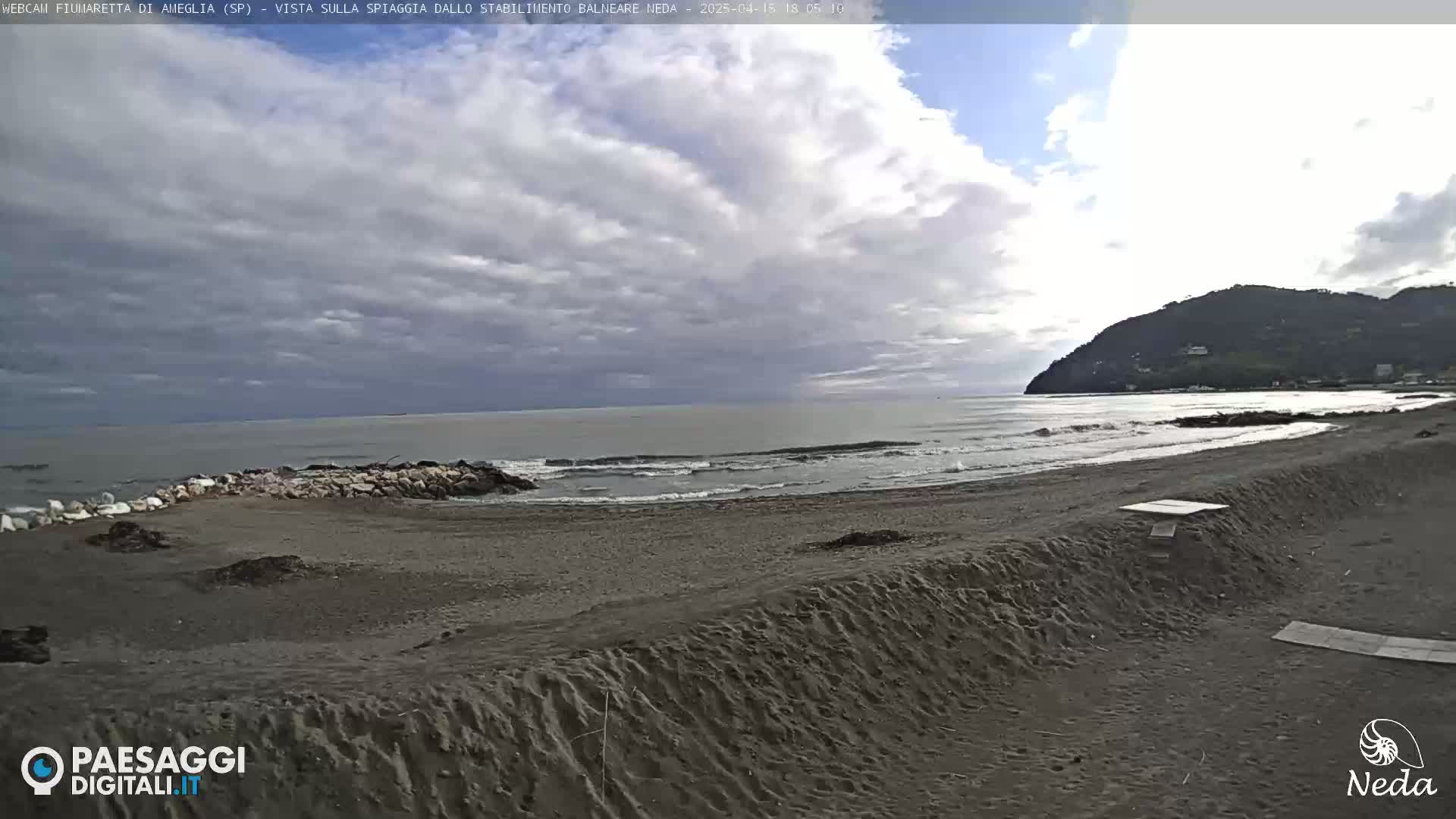 A mostly cloudy day at a sandy beach with small waves lapping the shore, backed by a hill.