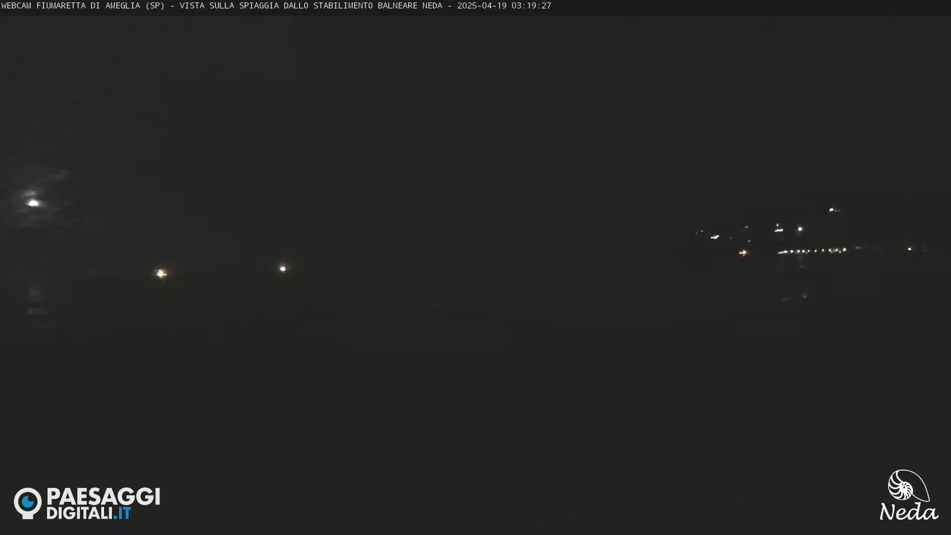 A nighttime, dark view of a distant, faintly lit shoreline under a mostly cloudy sky, with a bright moon visible.