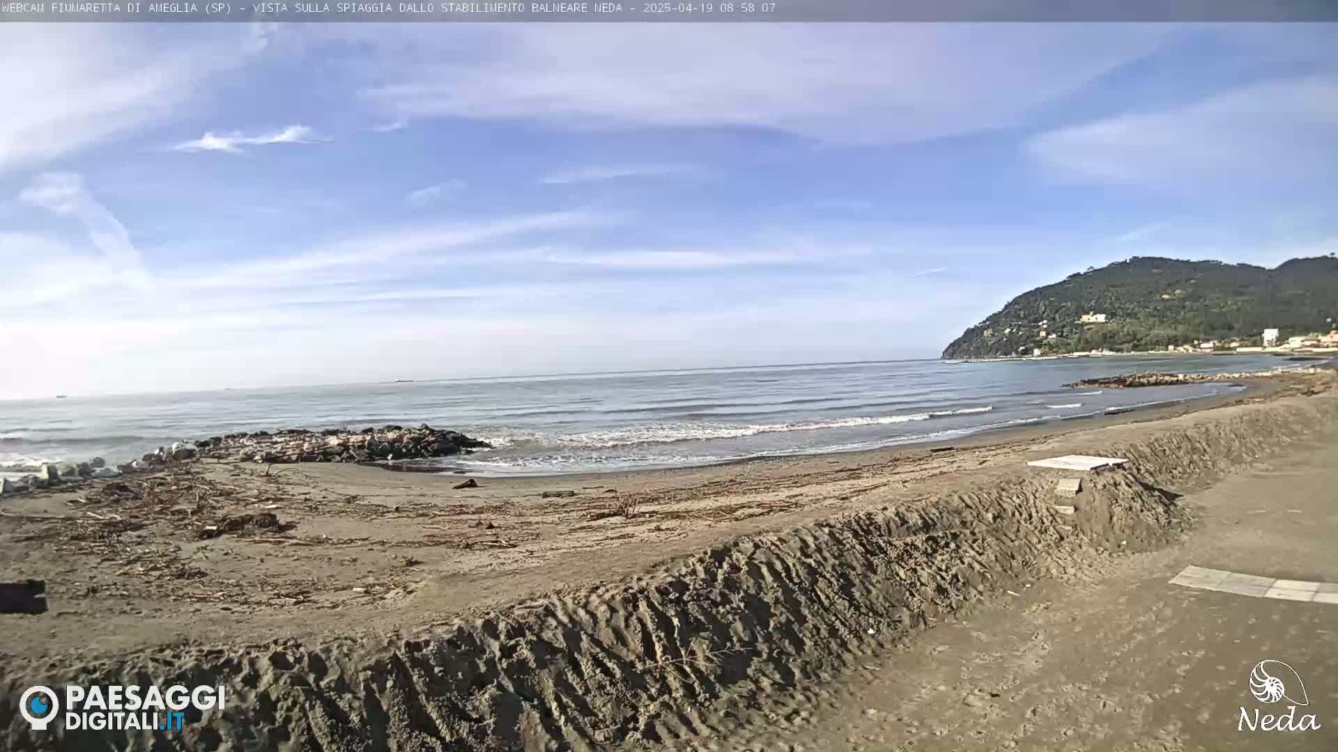 A sandy beach with small waves and a partly cloudy sky shows a rocky area at the water's edge and a hill in the distance.